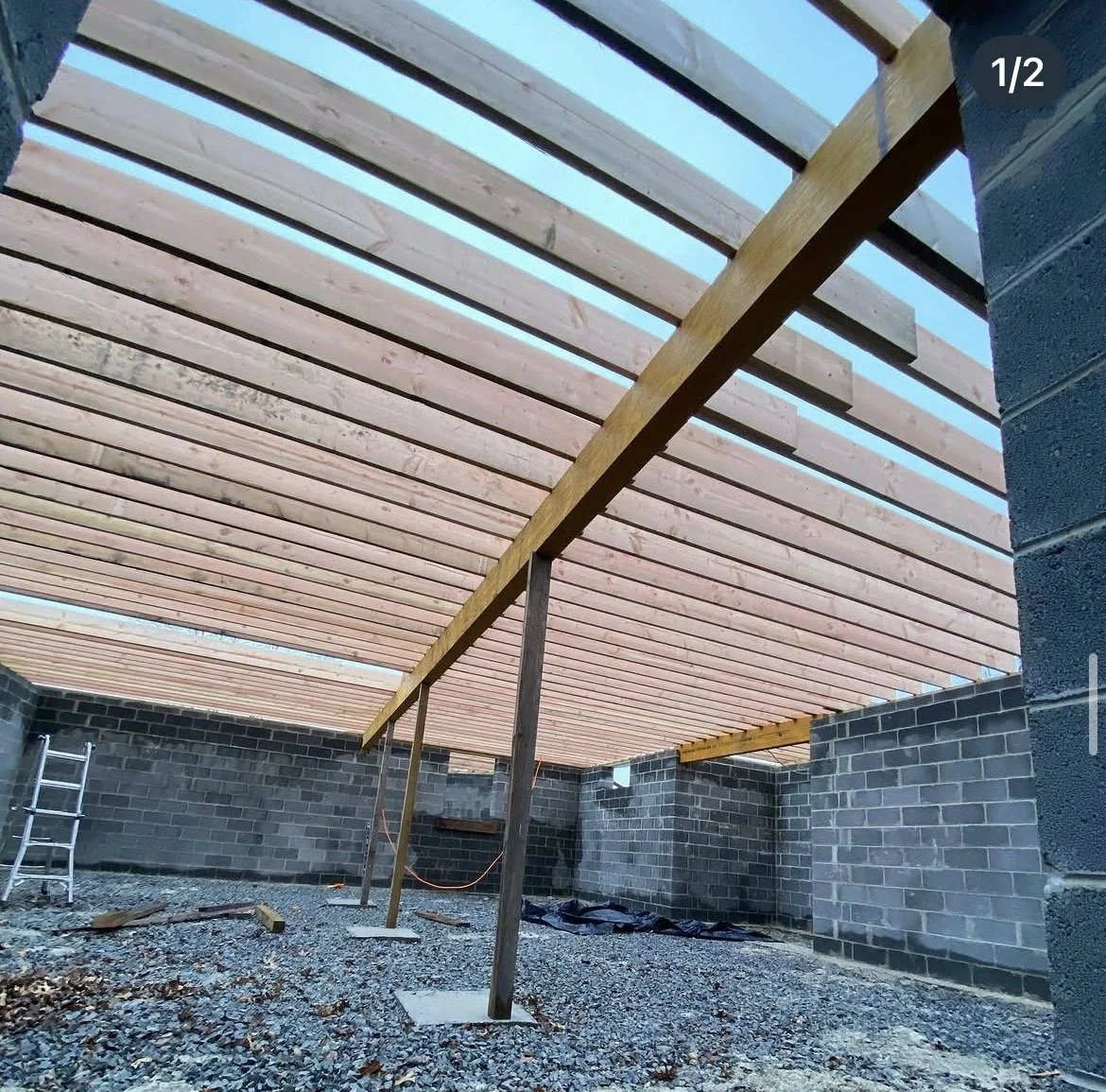 Construction site showing a wooden slatted roof structure supported by temporary wooden and metal posts, with brick walls and gravel ground.