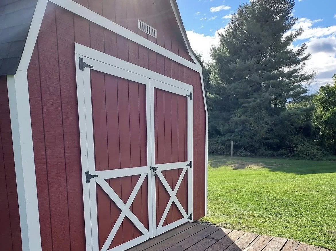 A red wooden barn with white trim, double doors, and a small ventilation window near the roof, set outdoors on a sunny day with green grass and tall trees in the background.