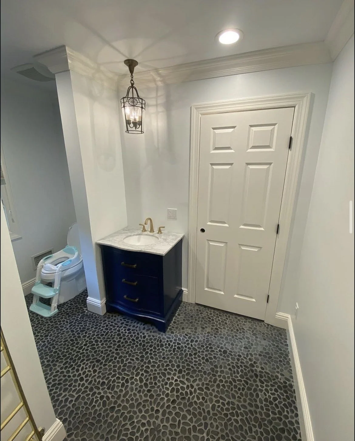 Small bathroom with black pebble tile floor, white walls, a white six-panel door, a small navy blue vanity with a marble countertop and gold faucet, a hanging lantern light fixture, and a white laundry basket next to a wall vent.