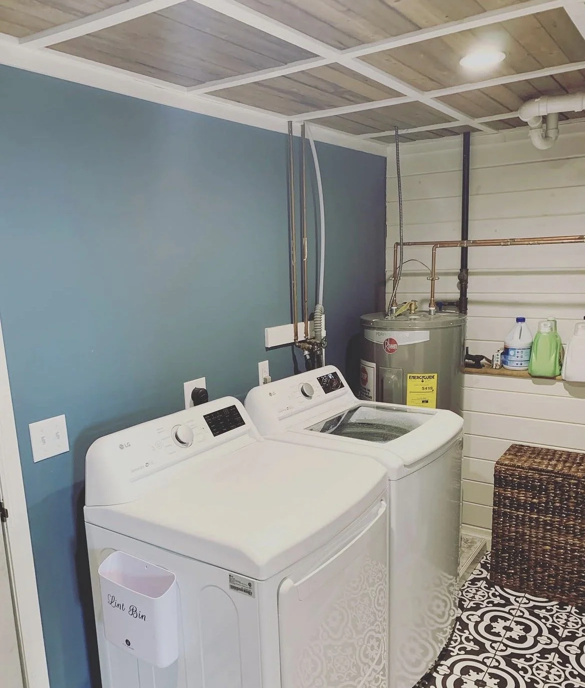 A laundry room featuring a white washing machine and dryer, a water heater, shelves with cleaning supplies, a blue accent wall, a wicker laundry basket, and patterned floor tiles.