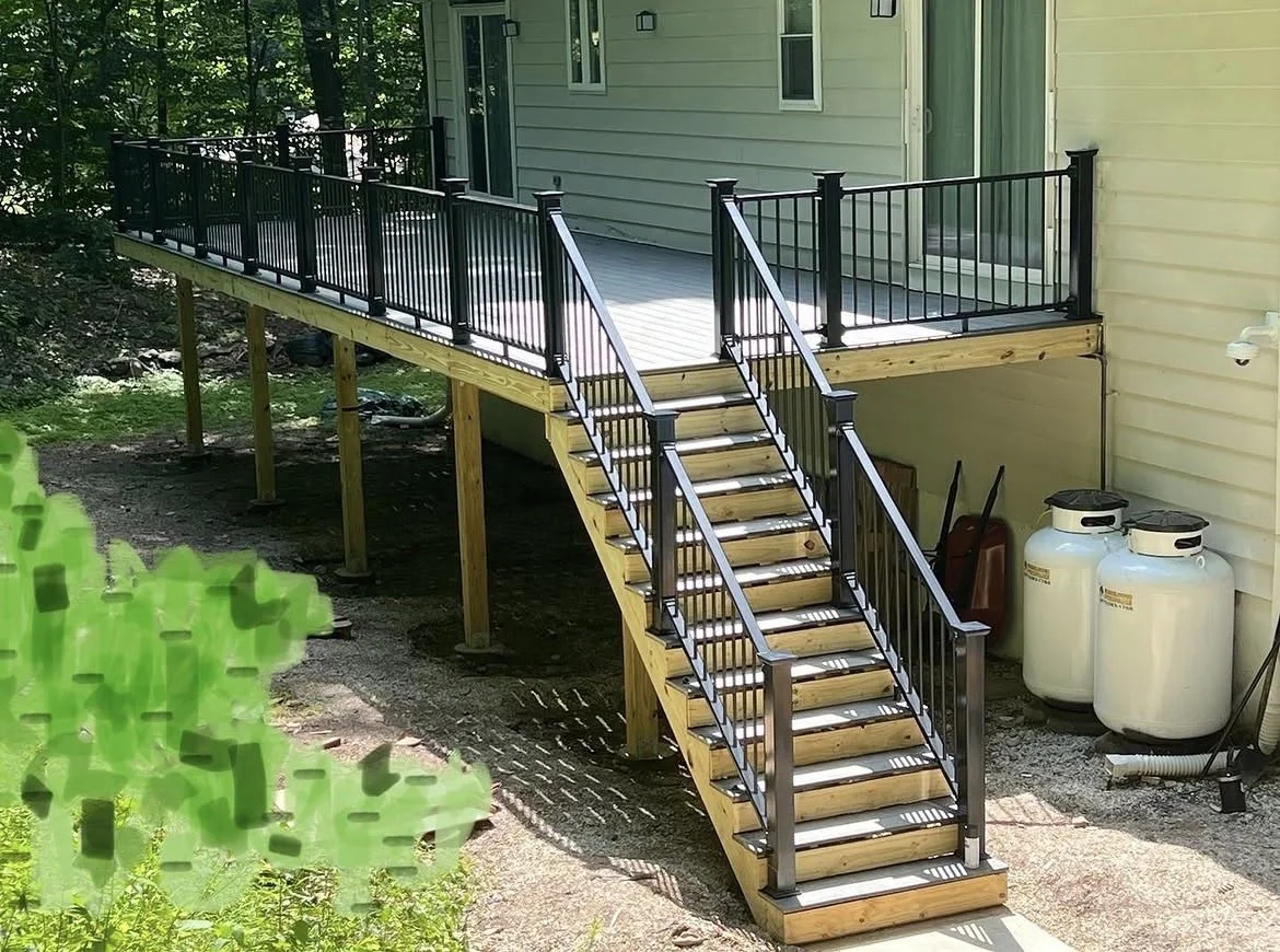 A newly built wooden deck with black metal railings attached to the back of a house, accessed by a staircase with matching black metal railing, with propane tanks and garden tools underneath.