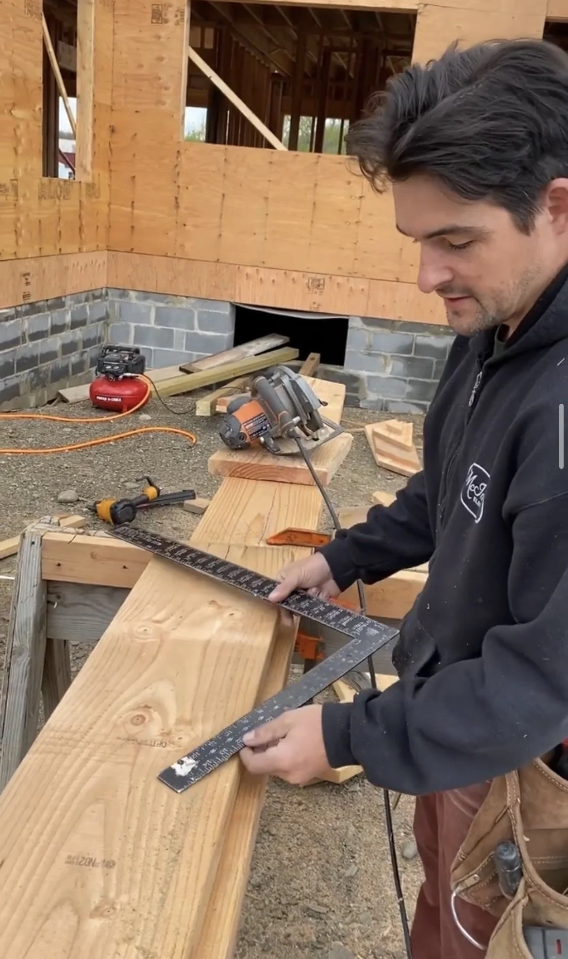 A man working on a construction site, using a level and a measuring tape on a wooden beam, with tools and building materials around him, and an unfinished building in the background.