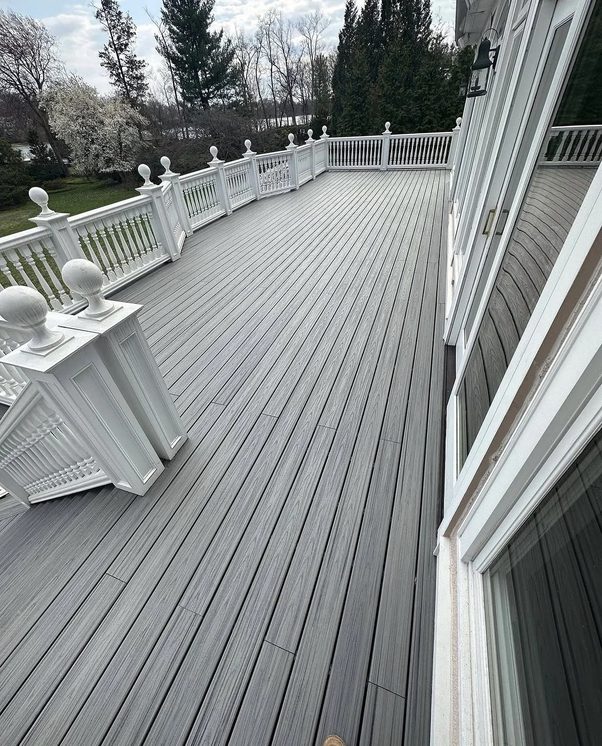Empty gray wooden deck with white railing, adjacent to a house with sliding glass doors, overlooking trees and a cloudy sky.