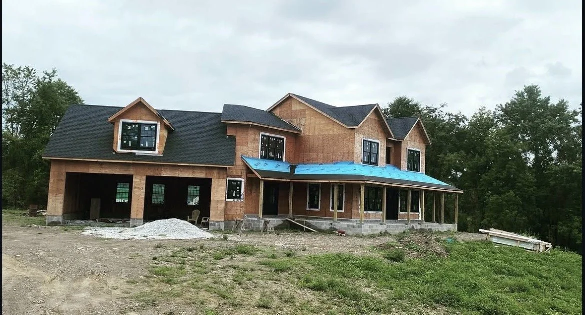 A two-story house under construction with beige brick exterior, black roof, and multiple windows, including some dormer windows, on a grassy lot with trees in the background.