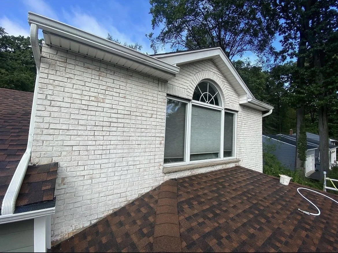 View of a house's exterior wall with a large arched window and white brick siding, attached to a roof with brown shingles, surrounded by trees.