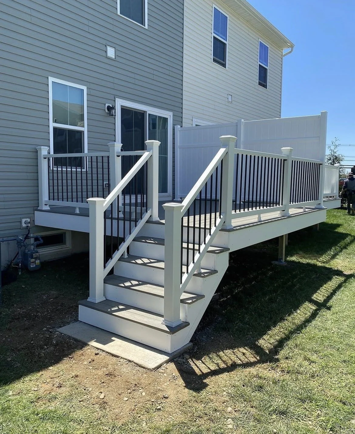White elevated deck attached to a house, with a staircase leading down to the yard, surrounded by a white and black railing, under a clear blue sky.