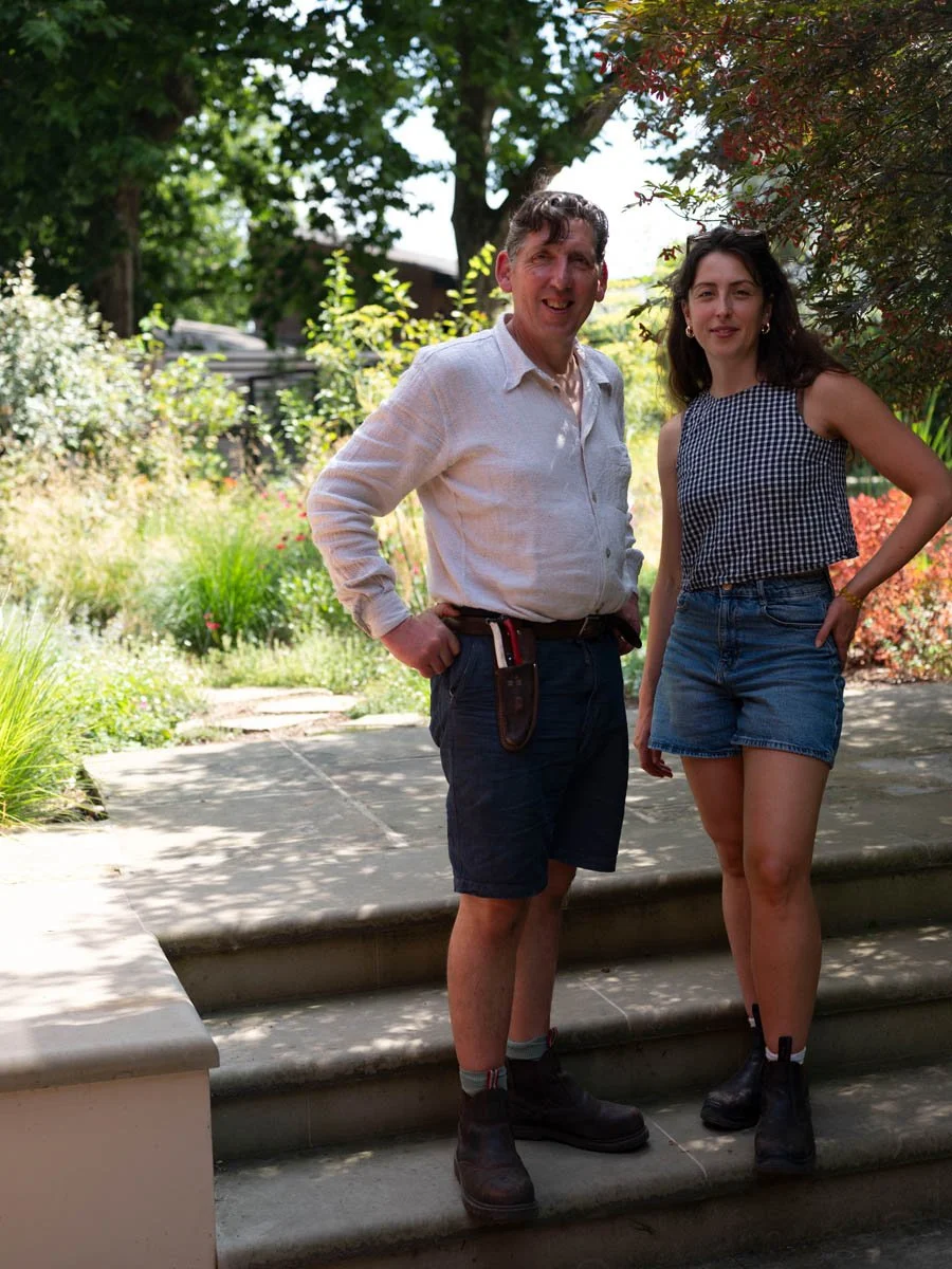 A man and a woman standing outdoors on a stone step in a garden with trees and plants, both smiling at the camera.