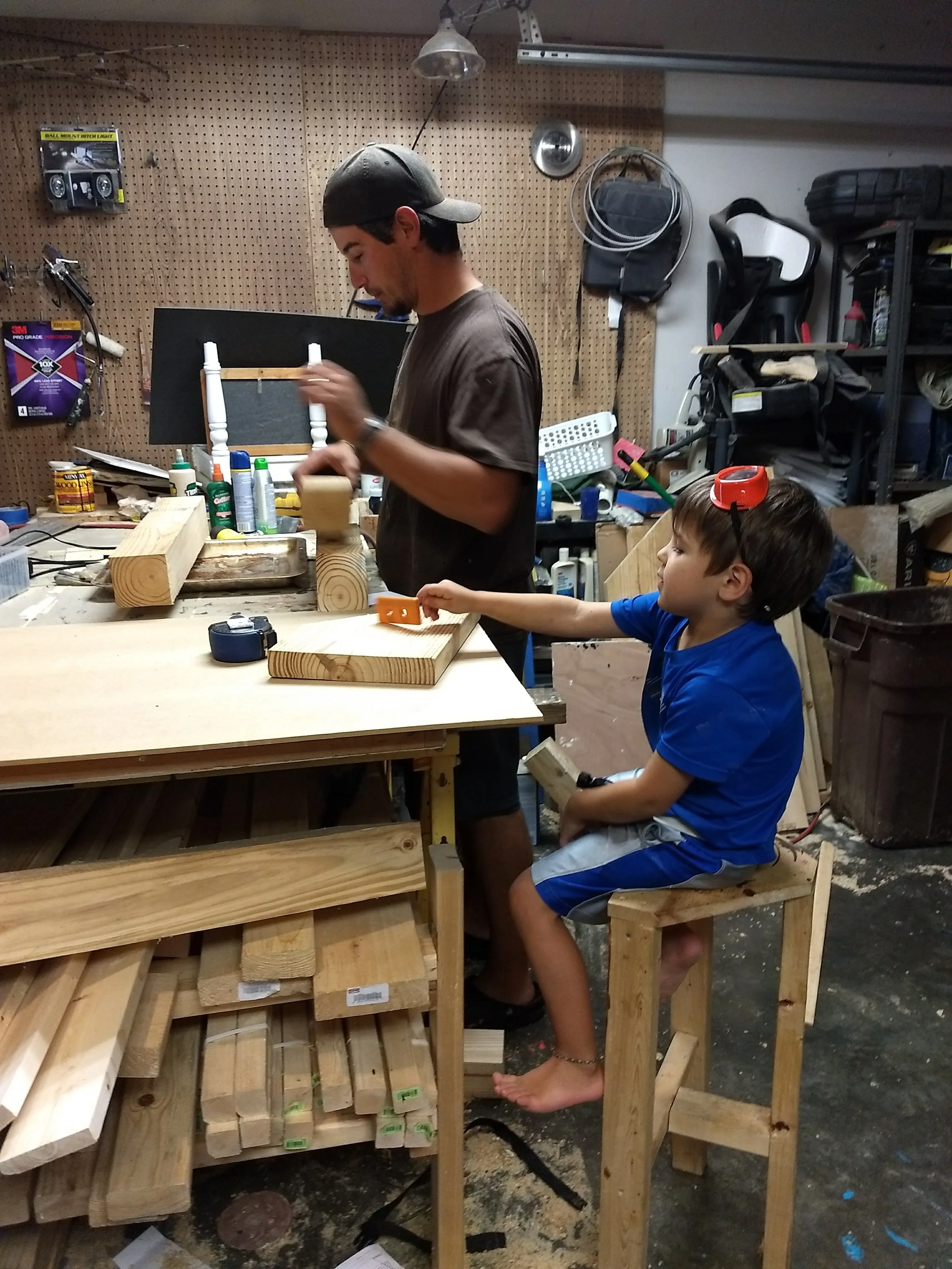Two people, a young man and a boy, working on wood projects in a workshop. The young man is sanding wood and wearing a backwards cap, while the boy, sitting on a chair, is holding a piece of wood and looking at it. The workshop has tools, supplies, and wooden pieces on the workbench and shelves.
