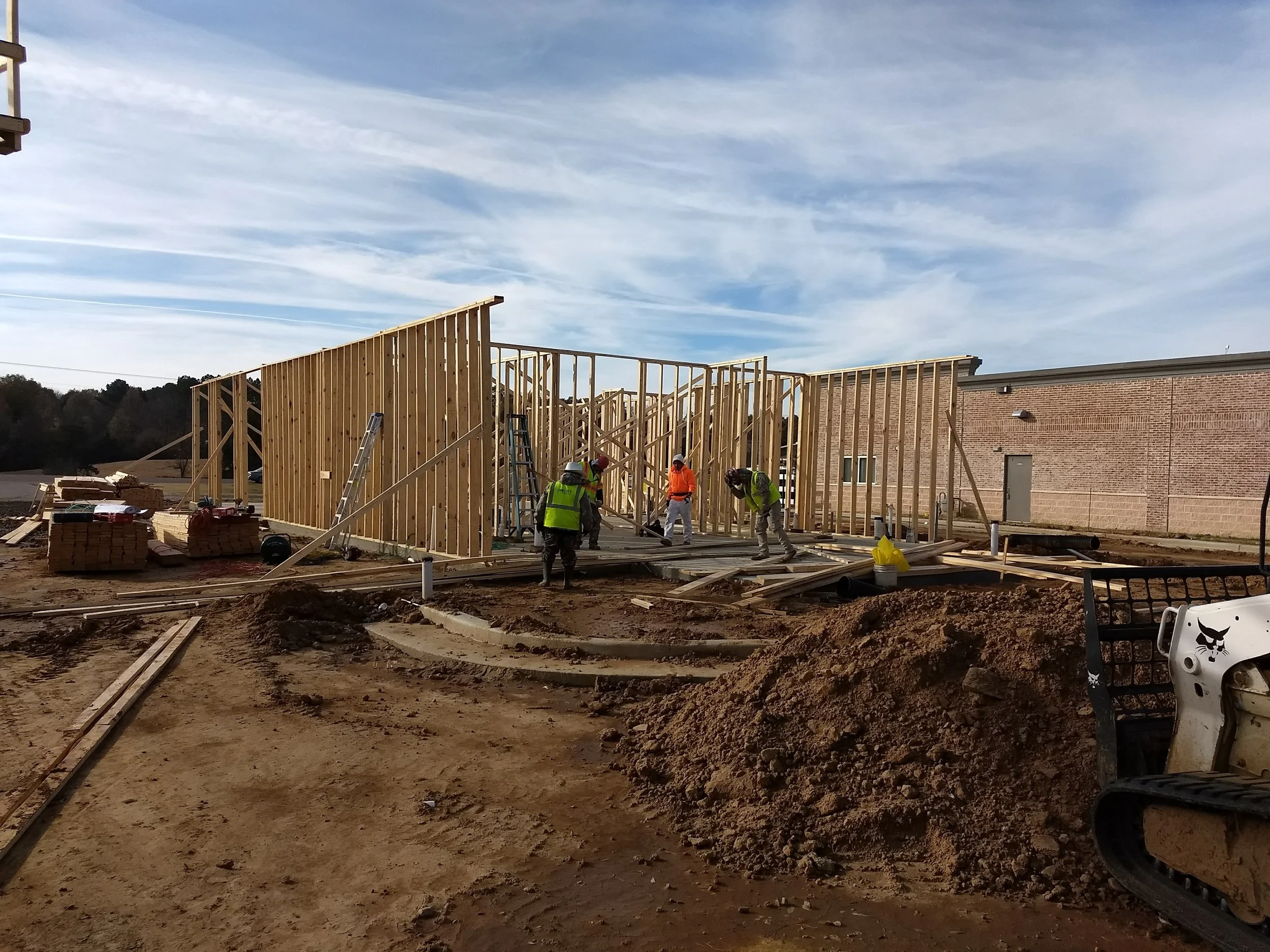 Construction site with workers building a wooden frame structure, with a brick building in the background and a partly cloudy sky above.