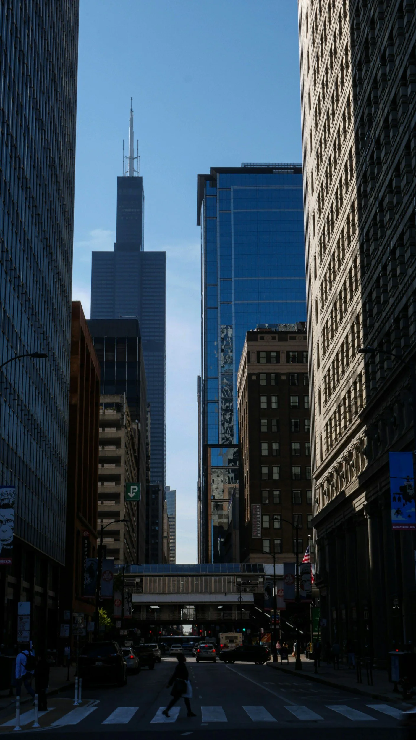 A city street scene with tall skyscrapers on either side and a clear blue sky above. Pedestrians are crossing the street at a crosswalk, and cars are driving down the road. Downtown buildings, some with reflective glass facades, line the street.