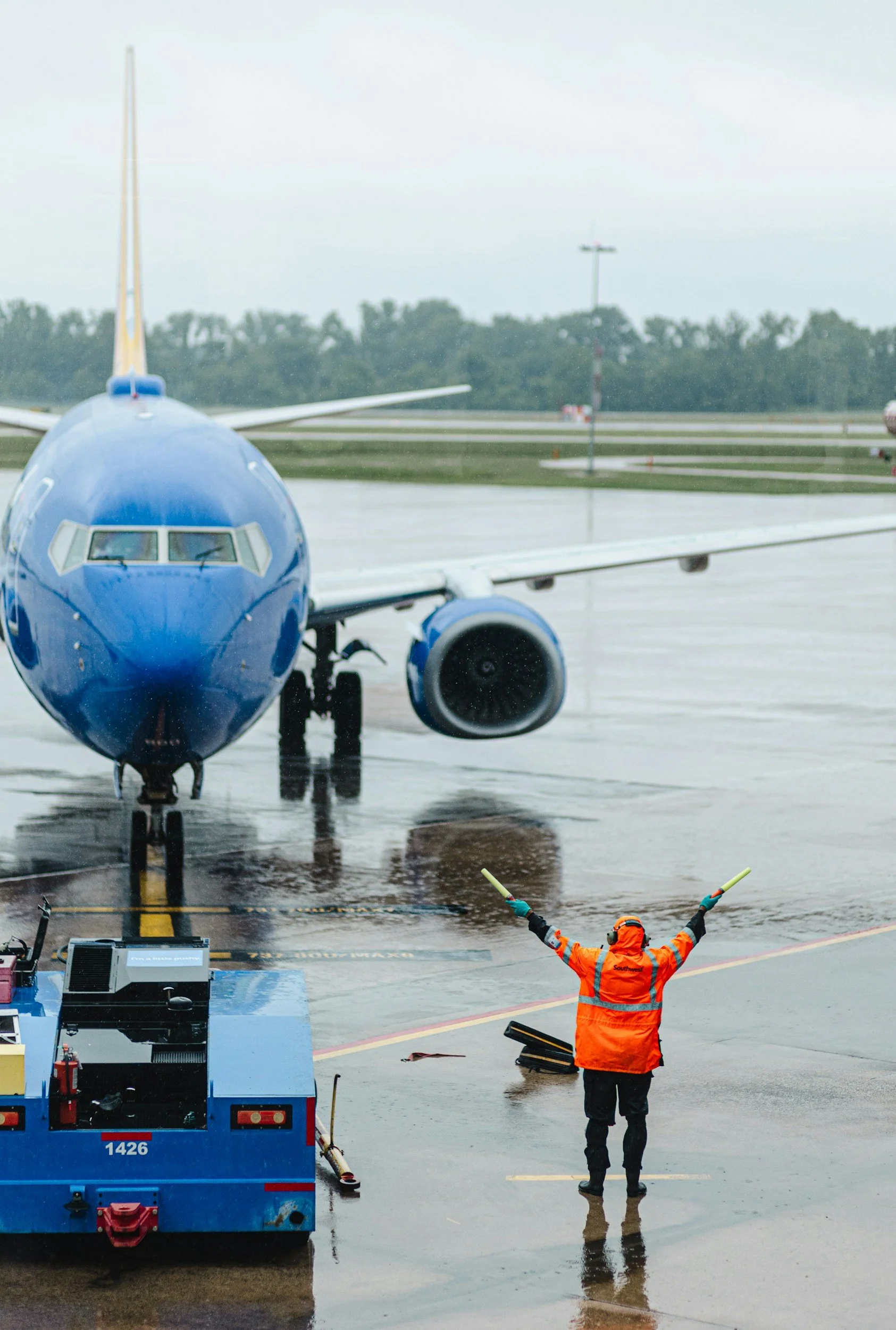 Ground crew member in orange safety jacket guiding a blue airplane on wet tarmac at an airport during overcast weather.