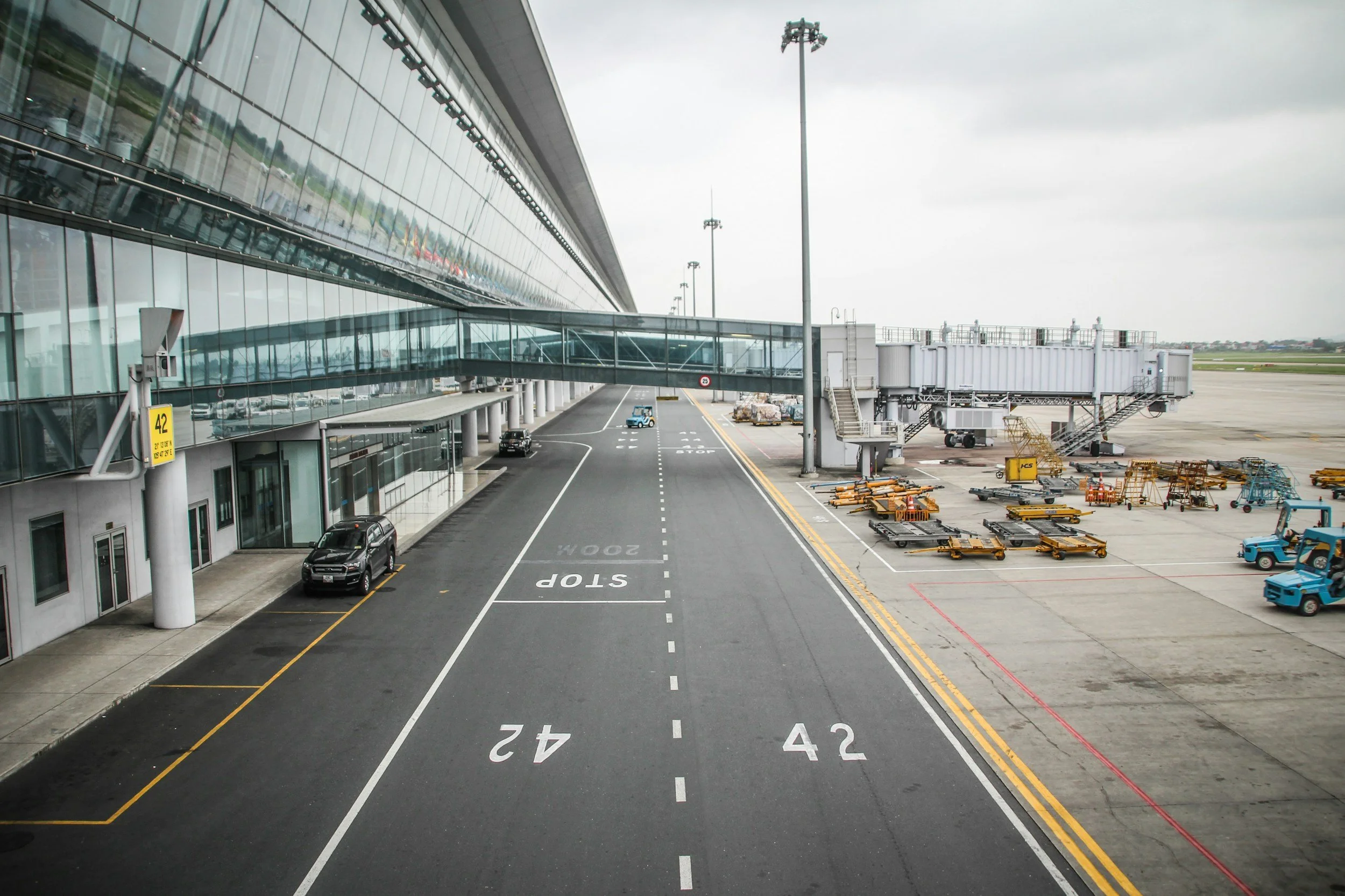 View of an airport tarmac with a modern glass terminal building and a jet bridge extending to an aircraft not visible in the image. The tarmac has marked parking spots with numbers, and there are various airport ground support vehicles and equipment, including baggage carts and service trucks. The sky is cloudy.