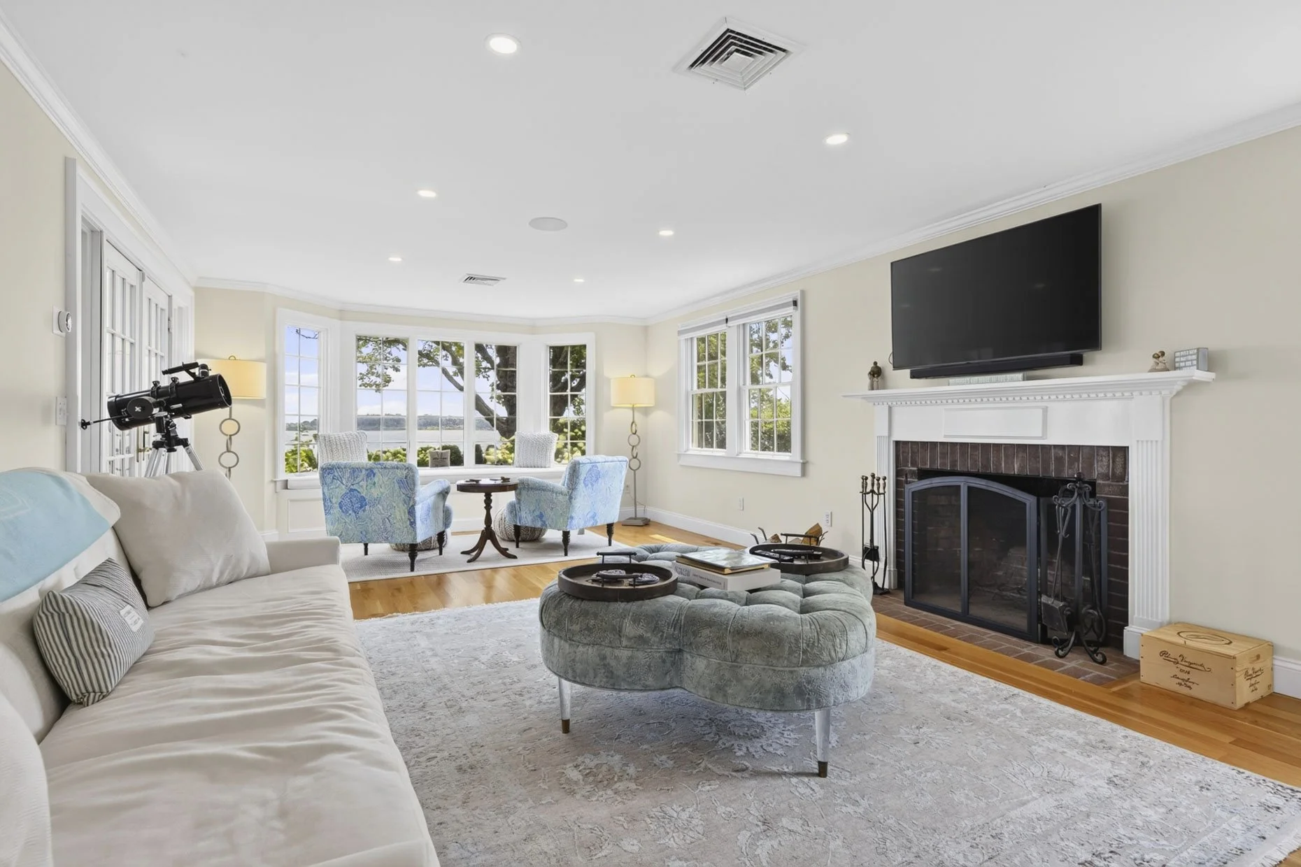 Bright living room with large bay windows overlooking water and trees, white walls, hardwood floors, a cream sofa with pillows, a green tufted ottoman with trays, a fireplace with a mounted TV, a telescope, and blue armchairs around a small table.