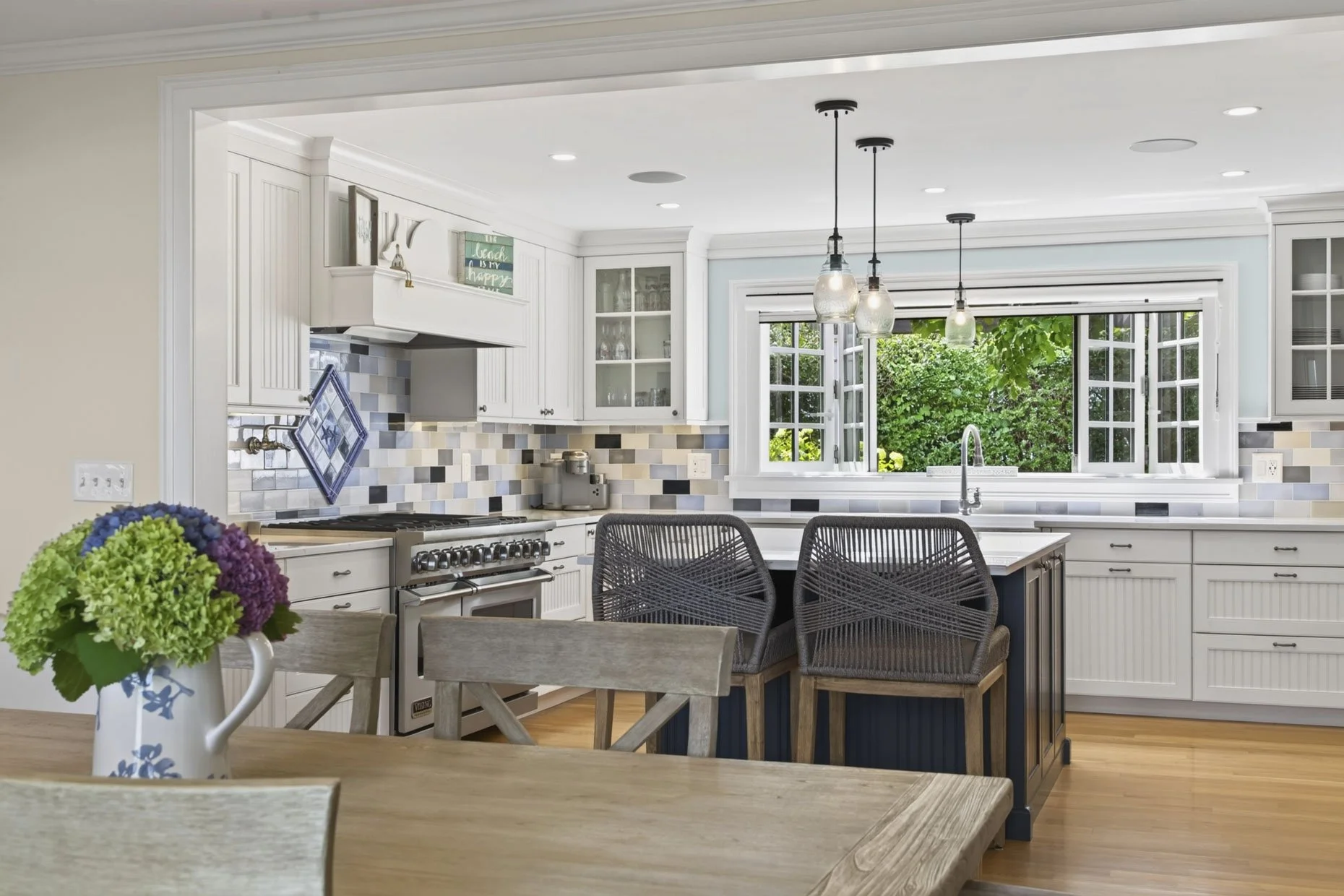 A bright and modern kitchen with white cabinetry, a mosaic tile backsplash, a window overlooking greenery, and a kitchen island with seating. There is a flower arrangement on the dining table in the foreground.
