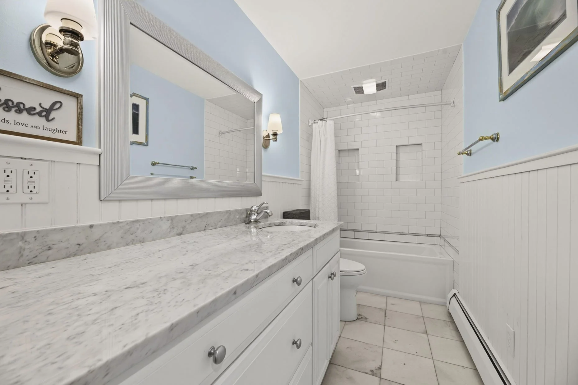 Bathroom with light blue walls, white wainscoting, a marble countertop, large mirror, wall-mounted light fixtures, and a shower with white subway tiles and built-in shelves.