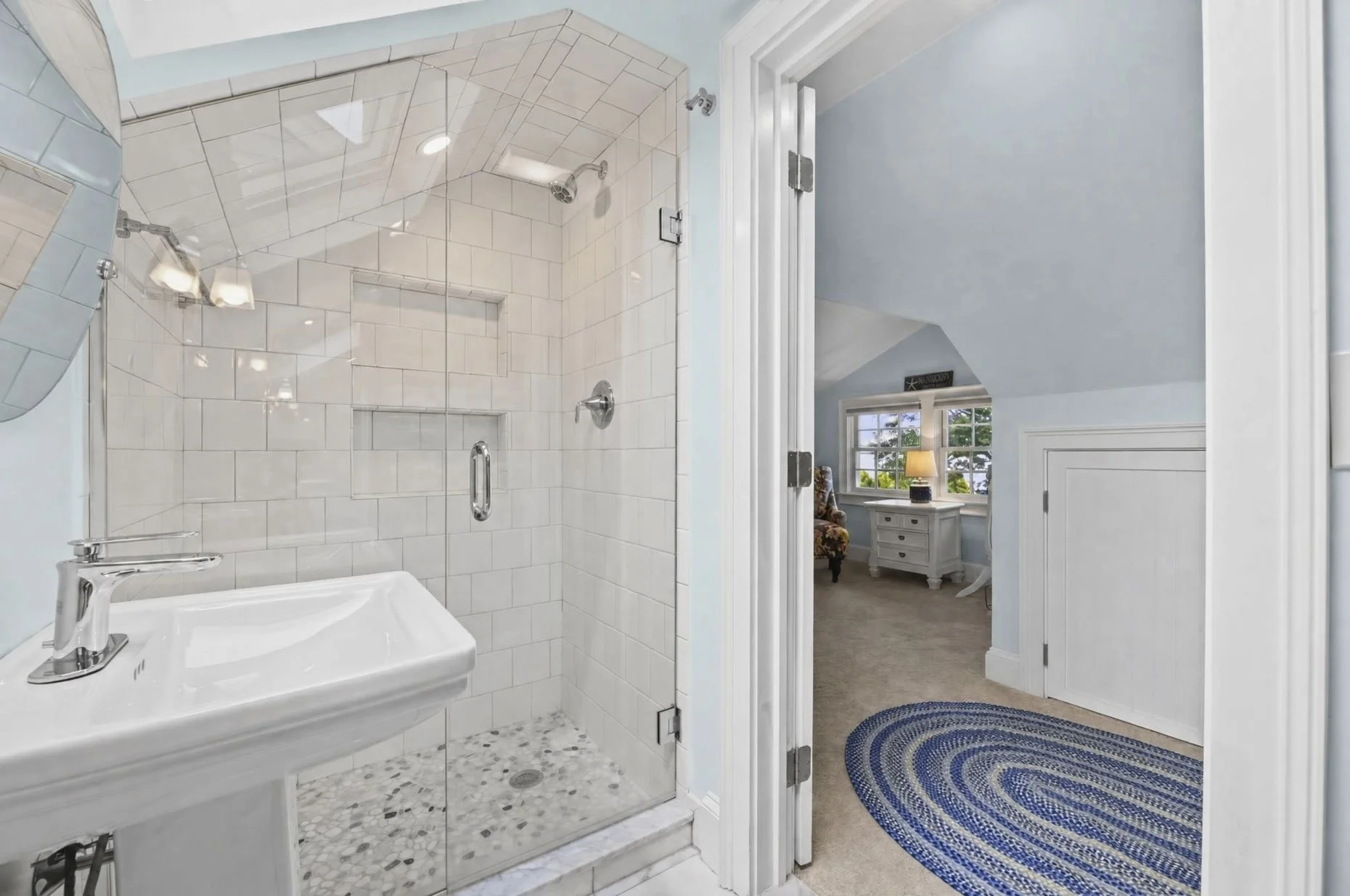 Bathroom with a glass shower enclosure and white tiled walls, view into an adjacent room with a blue oval rug, a white dresser, a lamp, and a window with trees outside.