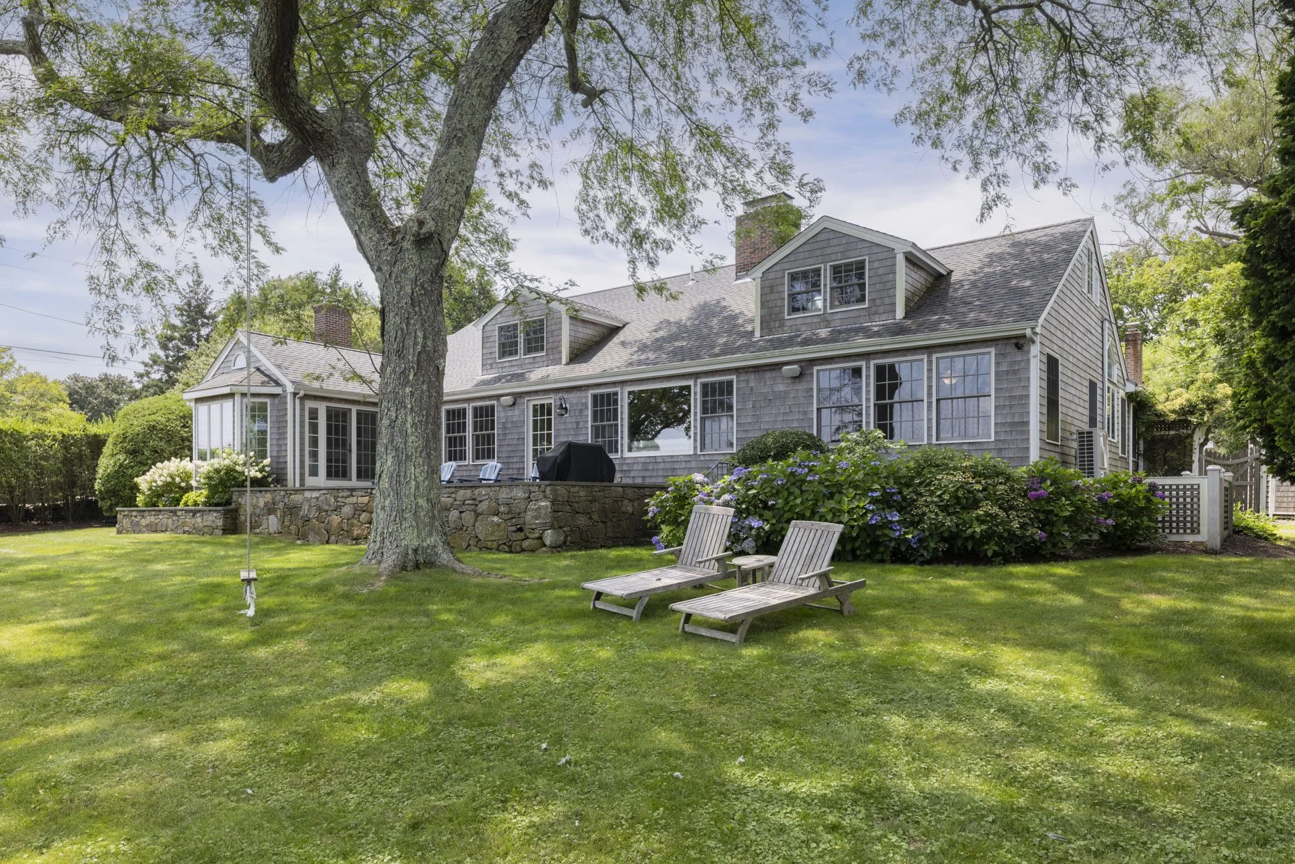 A backyard view of a large gray house with a stone patio, surrounded by green grass, bushes, and trees, with two wooden lounge chairs in the foreground and a swing hanging from a tree.