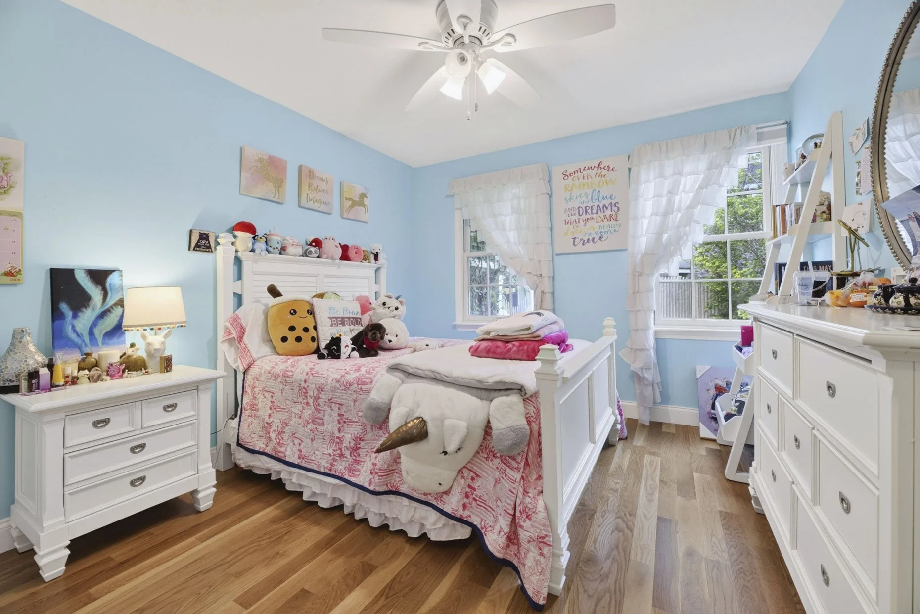 A bright blue bedroom with white furniture, a bed covered in pink and white bedding, plush toys on the bed, and a window with sunlight and white curtains.
