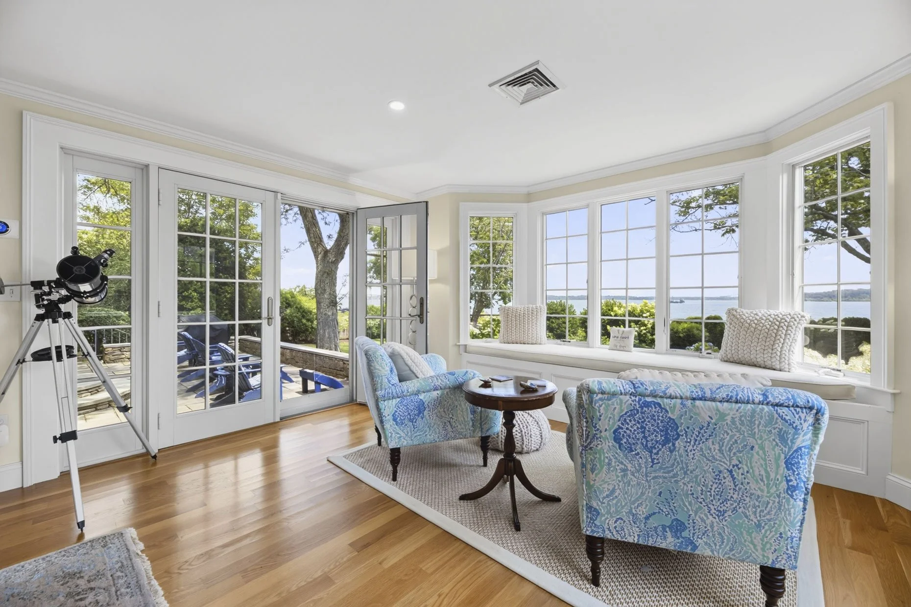 Living room with large bay windows overlooking water and trees, featuring blue patterned armchairs, a small round wooden table, a window seat with pillows, and a telescope aimed out the door to the patio.