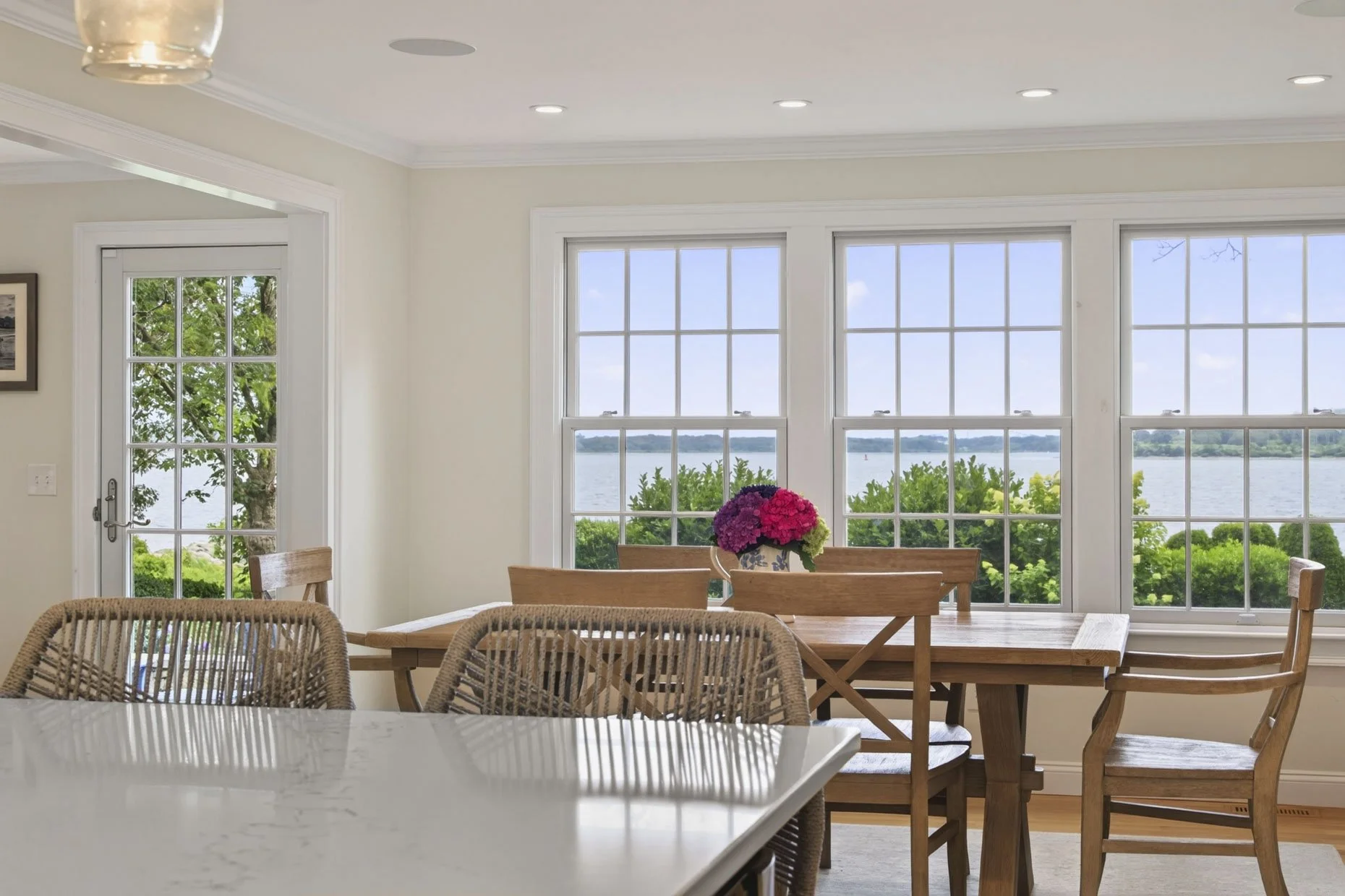 Dining area with a wooden table, floral centerpiece, large windows overlooking water and greenery, wicker chairs, and a door leading outside.