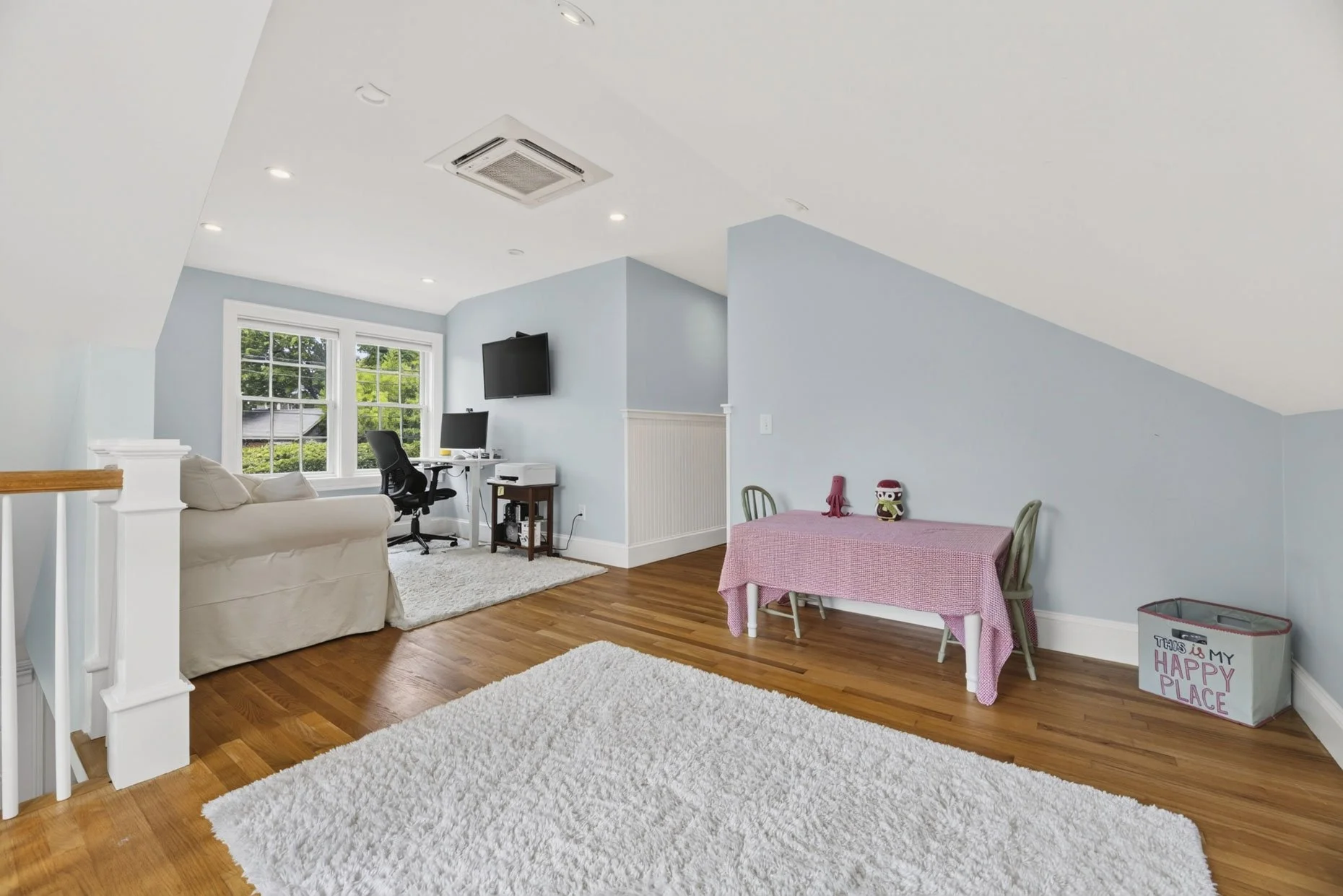 Living room with light blue walls, hardwood floors, large window with greenery outside, white couch, black office chair, small desk with computer, wall-mounted TV, pink table with two chairs and stuffed animals, white rug, laundry basket with 'This i
