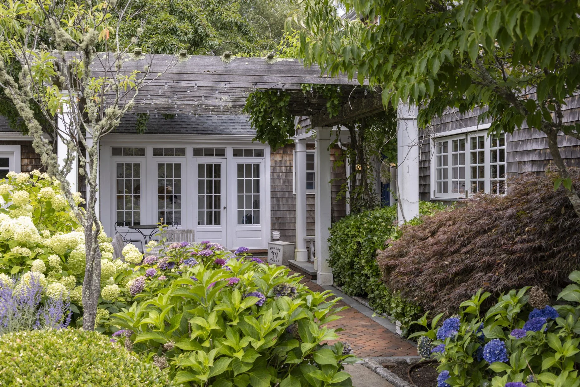 A charming garden with lush green foliage, colorful hydrangeas, and a brick pathway leading to a house with white-framed glass doors and windows, surrounded by trees and plants.
