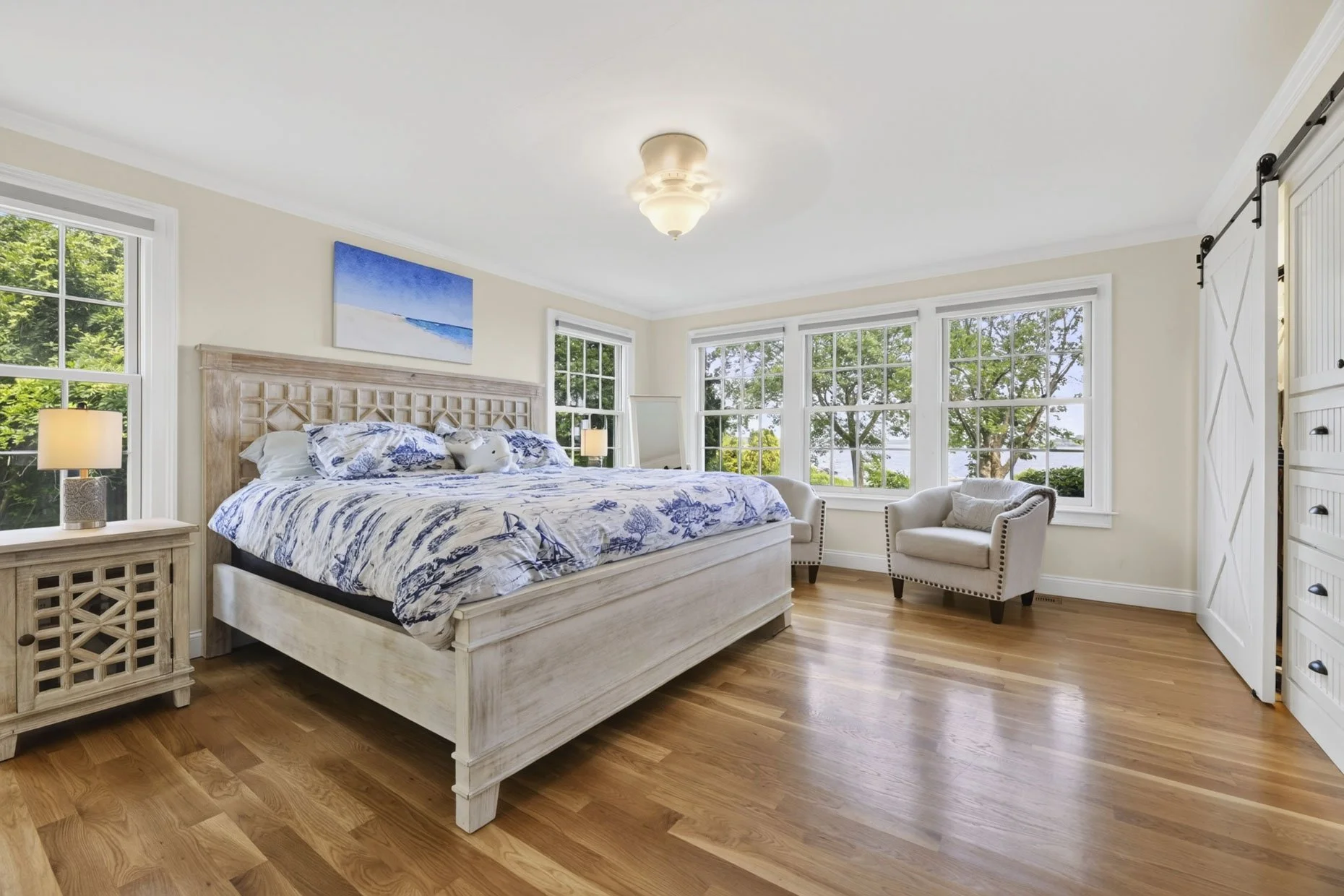 Bright bedroom with a white wooden bed, blue and white linens, a nightstand with a lamp, and large windows showing green trees outside, with a white armchair and barn door closet.