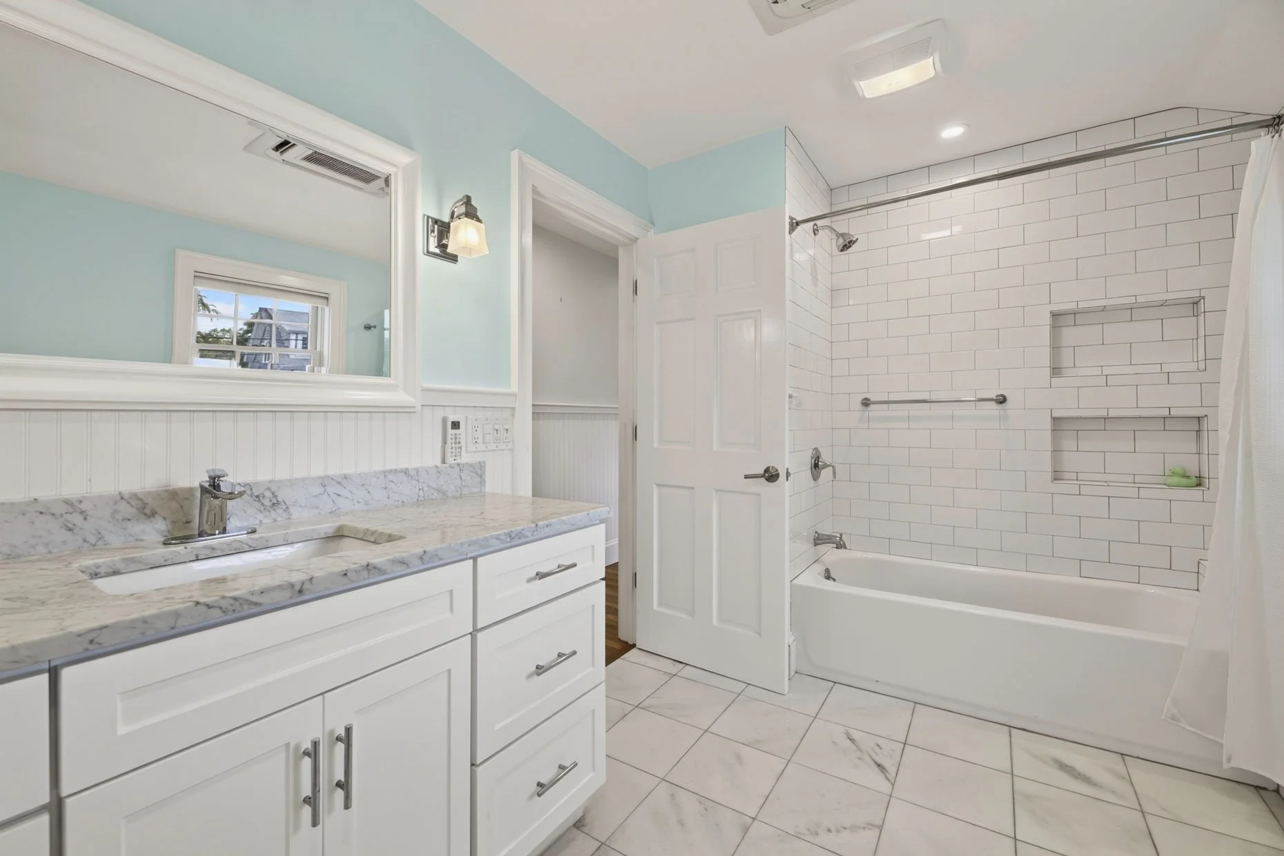 Bathroom with white vanity, marble countertop, and a mirror above. The shower area has white subway tiles and built-in shelves, with a showerhead and grab bars. There is a small window and ceiling lights.