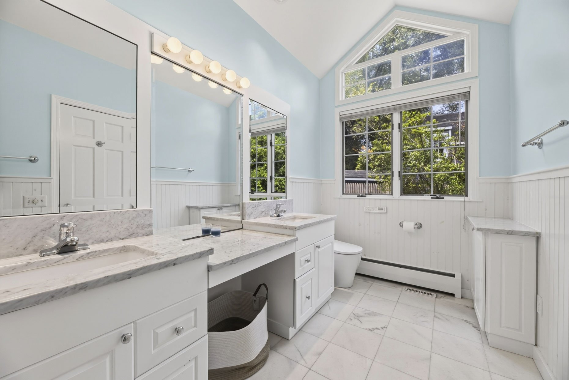 Bright bathroom with white cabinets, marble countertops, large window with trees outside, and a mirror above the sink with globe lighting.