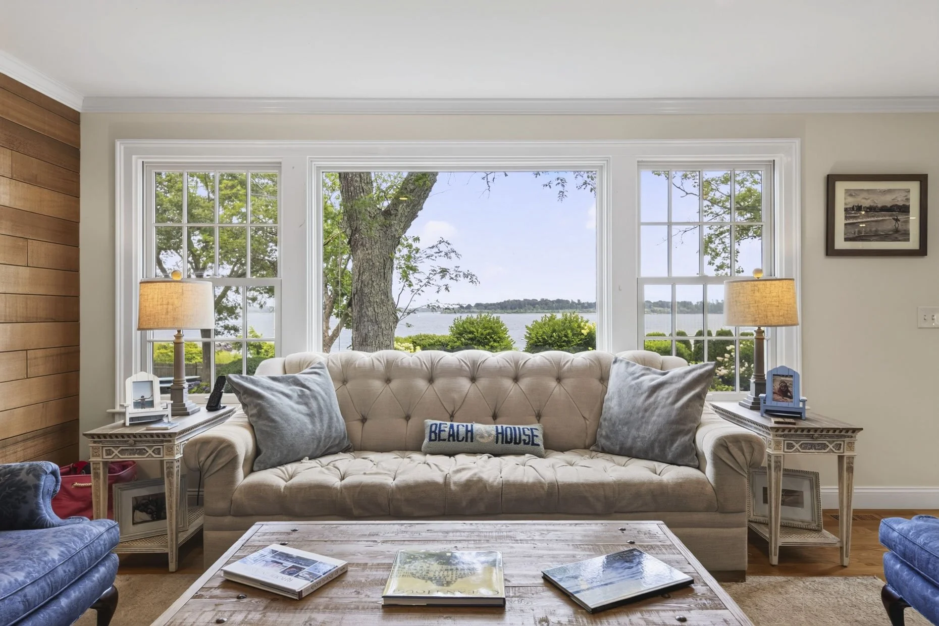 Living room with large window overlooking water and trees, beige tufted sofa with pillows, side tables with lamps, framed picture on wall, wooden coffee table with magazines.