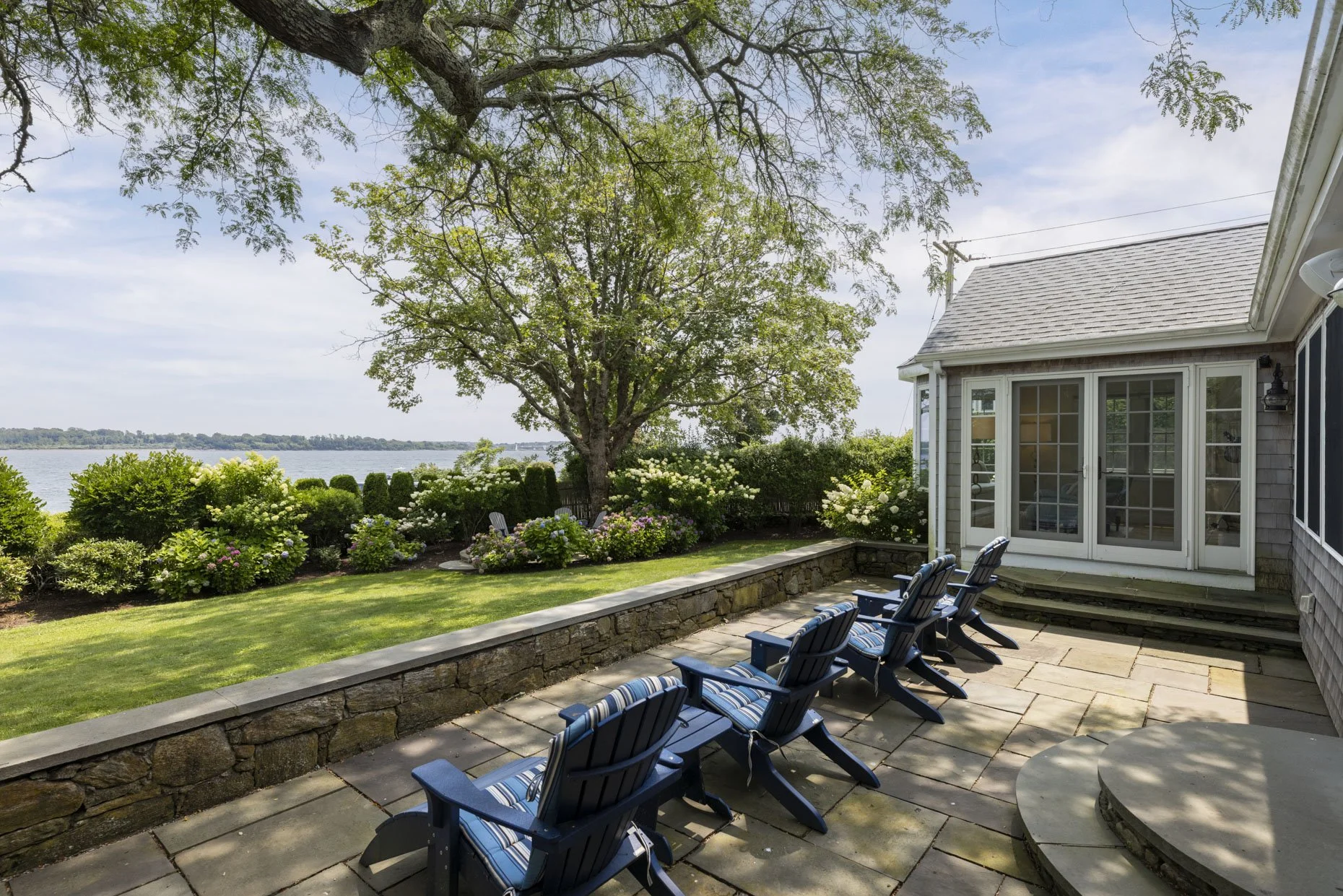 A backyard patio with Adirondack chairs facing a garden with blooming bushes and trees, overlooking a body of water with a distant shoreline.