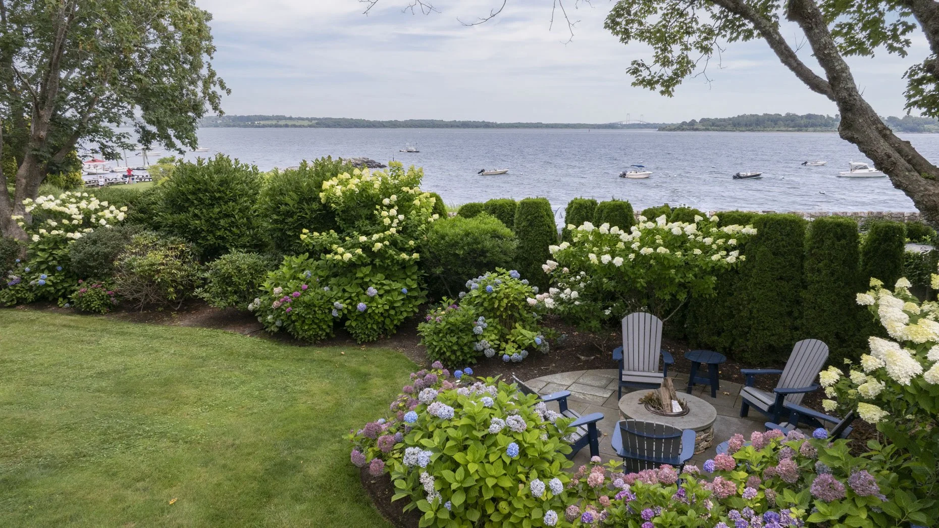 A scenic backyard garden with colorful hydrangea bushes, green hedges, and trees overlooking a body of water. There are boats on the water and a picnic area with chairs and a fire pit on a stone patio.