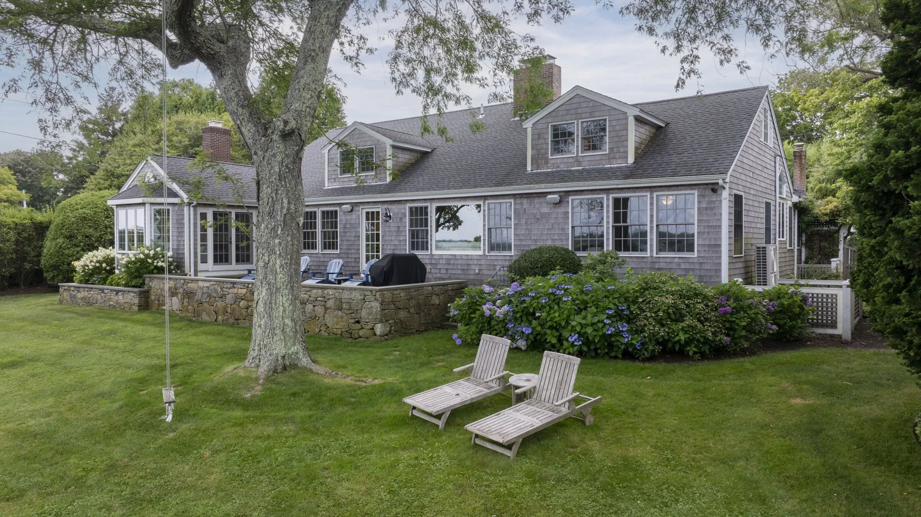 Large gray house with multiple windows and a stone patio in a lush green yard with trees, bushes, and flowers, with outdoor chairs and a grill.