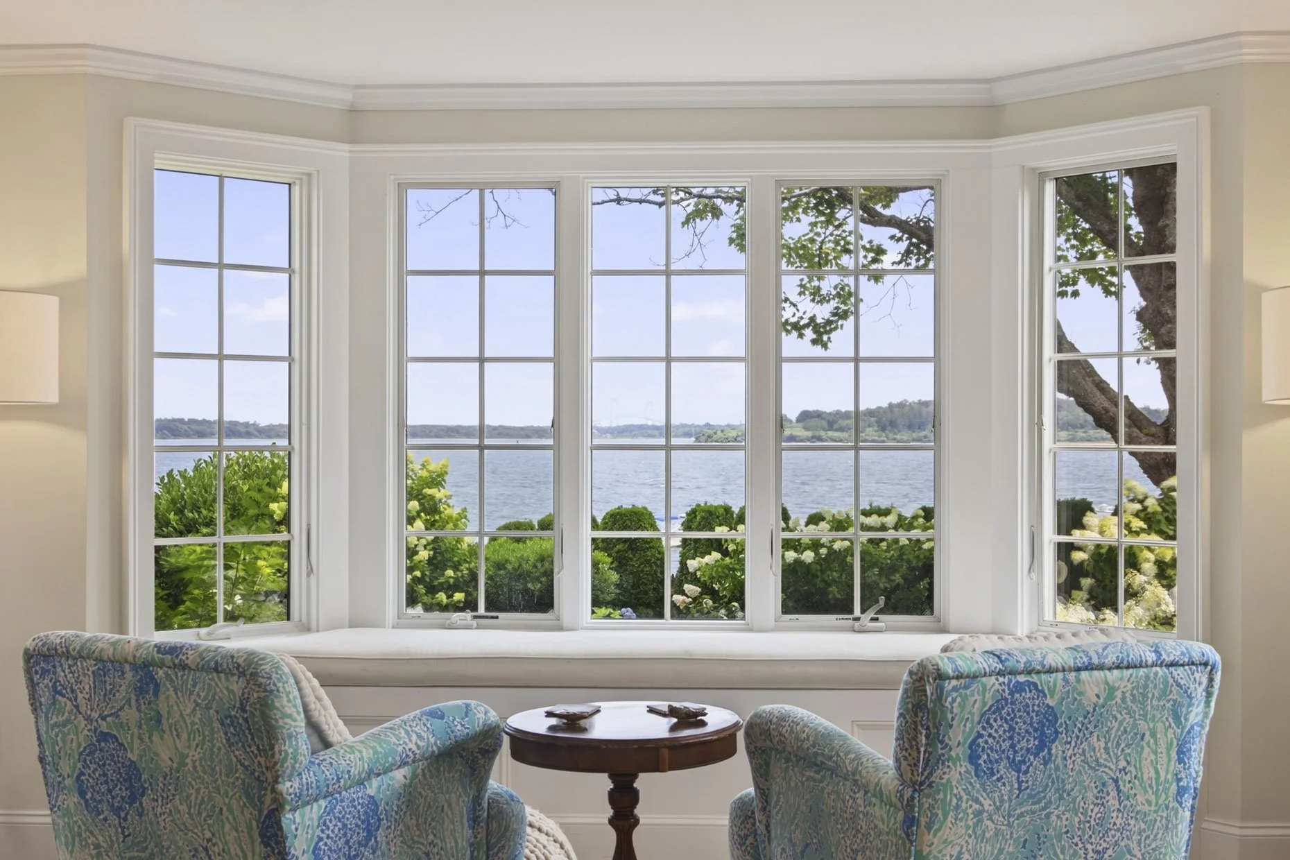 A cozy sitting area with two upholstered armchairs featuring a colorful blue, green, and beige pattern, a small round wooden table with decorative objects, and large bay windows showcasing a scenic view of a body of water, lush greenery, and a large 