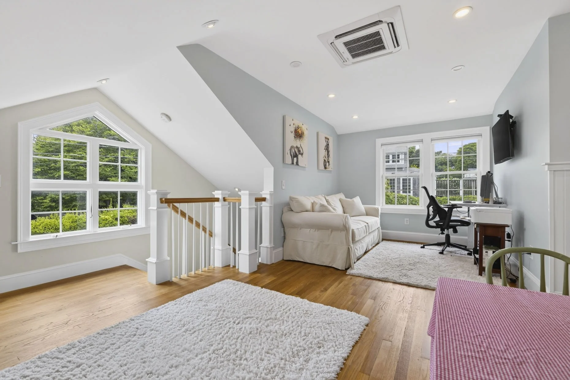 A cozy living space with a staircase, large windows showing greenery outside, a white couch, a small work desk with a computer, a black office chair, wall art, a white rug, and hardwood floors.