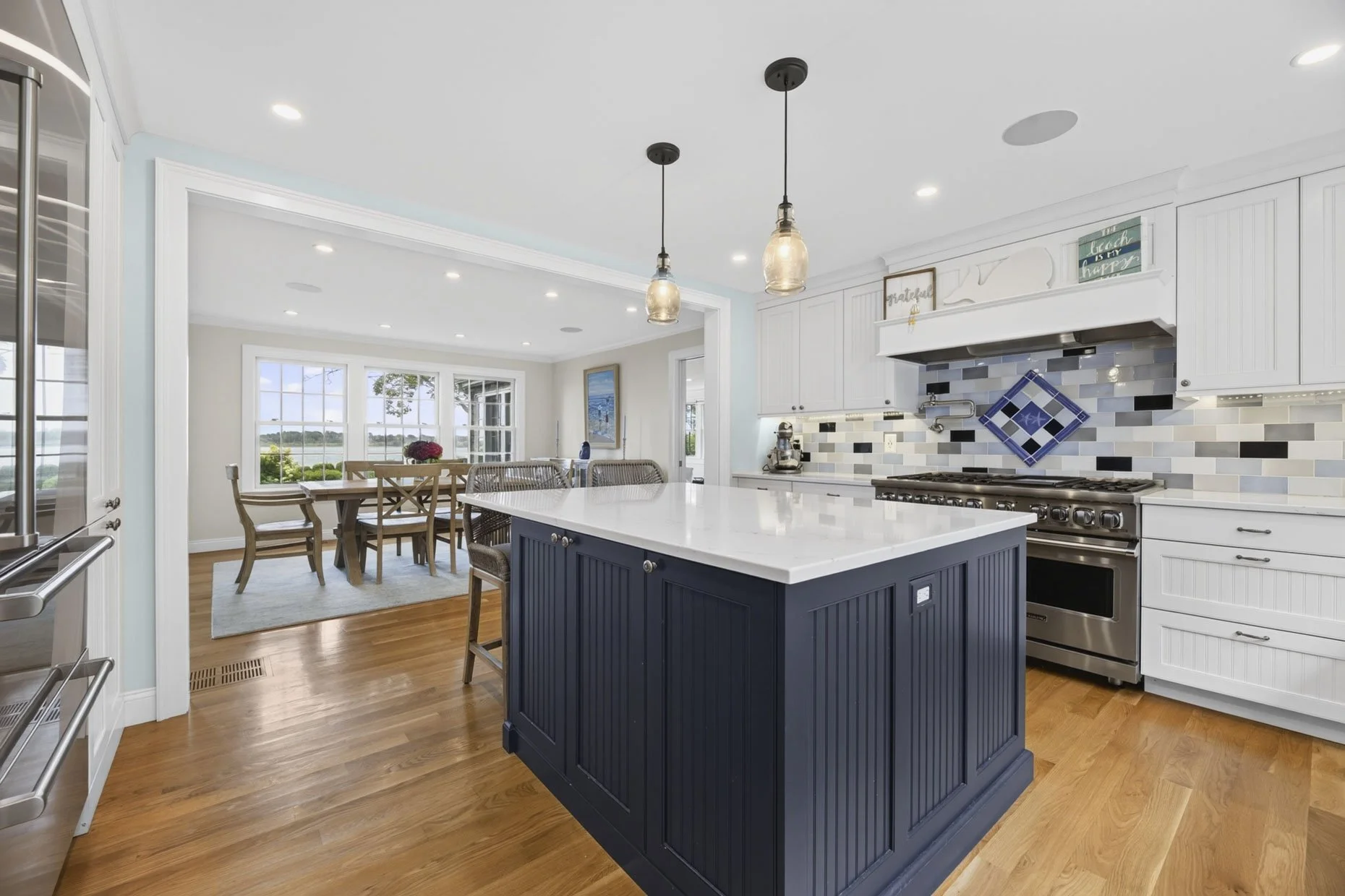 Modern kitchen with white cabinetry, a navy blue island, stainless steel appliances, a tiled backsplash, and pendant lights. Adjacent dining area with a wooden table and chairs near large windows.
