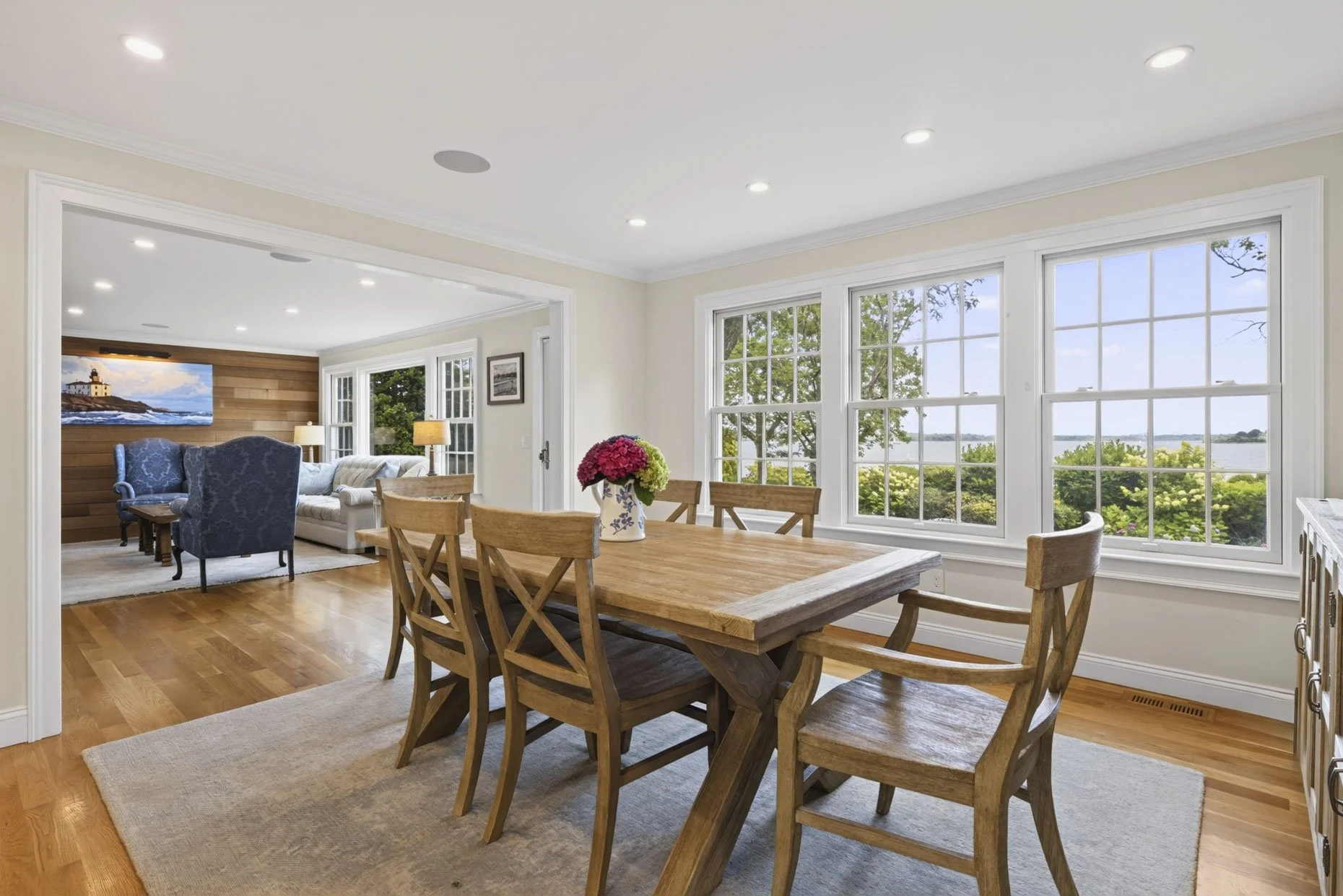 The image depicts a bright, open living and dining area with large windows overlooking a scenic water view and greenery. The dining area has a wooden table with matching chairs and a vase of pink and green hydrangeas. In the background, the living ar