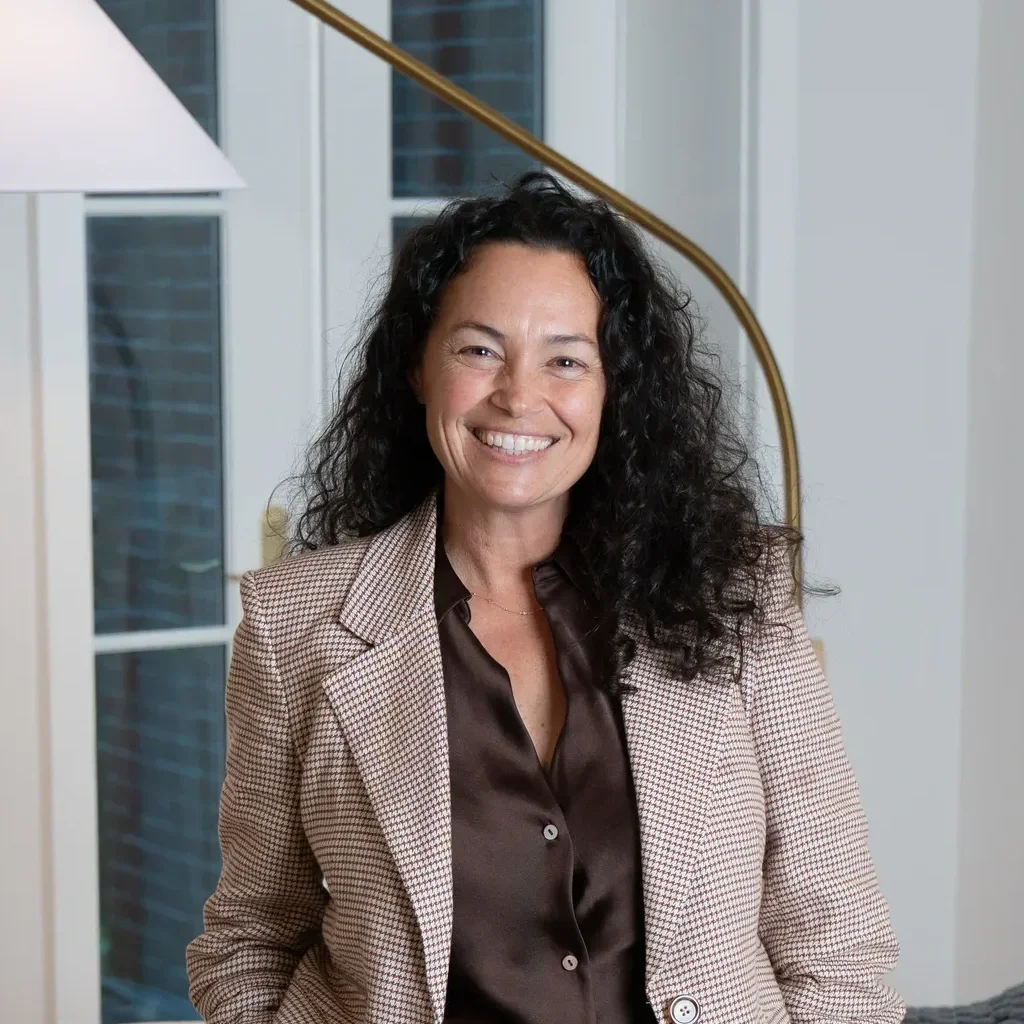 A woman with curly black hair smiling, wearing a light-colored blazer and dark blouse, standing indoors in front of a window and a staircase with a brass handrail.