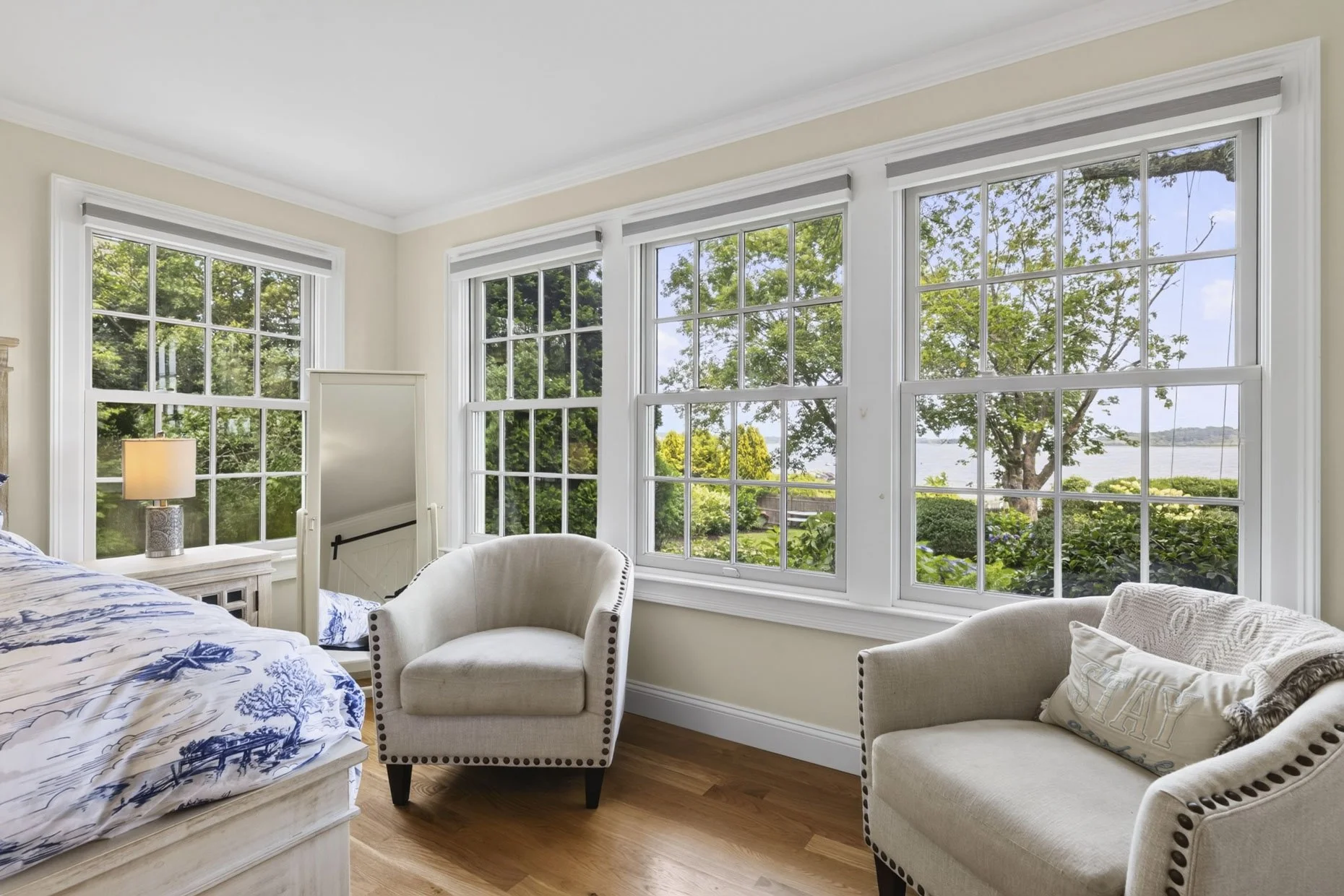 Living room with large windows showing trees and a view of the water outside, beige walls, wooden flooring, a white chair, a beige armchair with a pillow, and a side table with a lamp.
