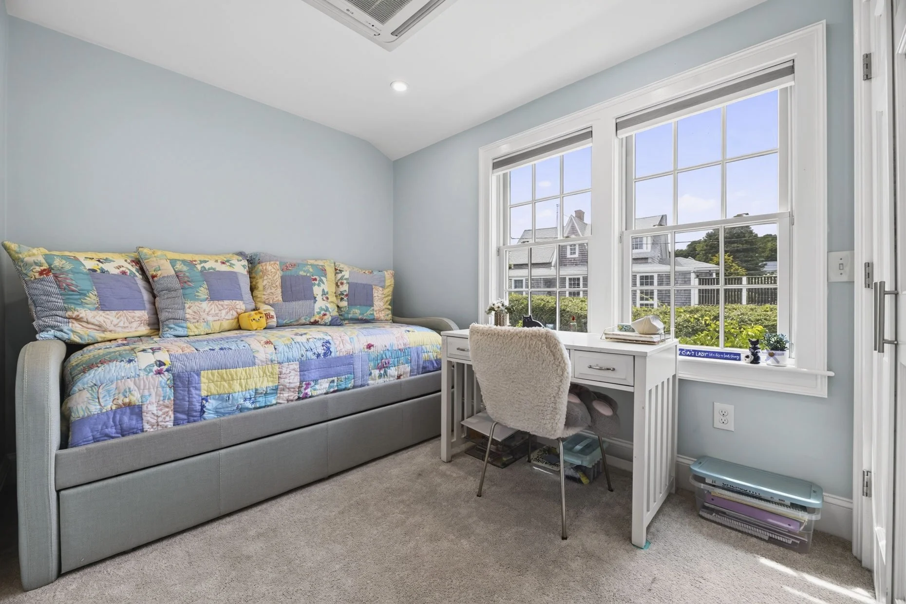 A cozy bedroom corner with a gray upholstered sofa covered with a colorful quilt and pillows, a white desk under two large windows with a view of neighboring houses, and a plush chair in front of the desk. Bottles of plants and decorative items sit o