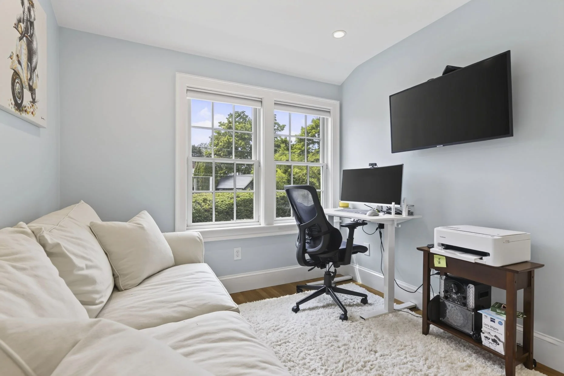 A bright home office with a white couch, two large windows showing greenery outside, a black ergonomic chair at a white desk with a computer monitor, and mounted flat-screen TV on light blue walls.