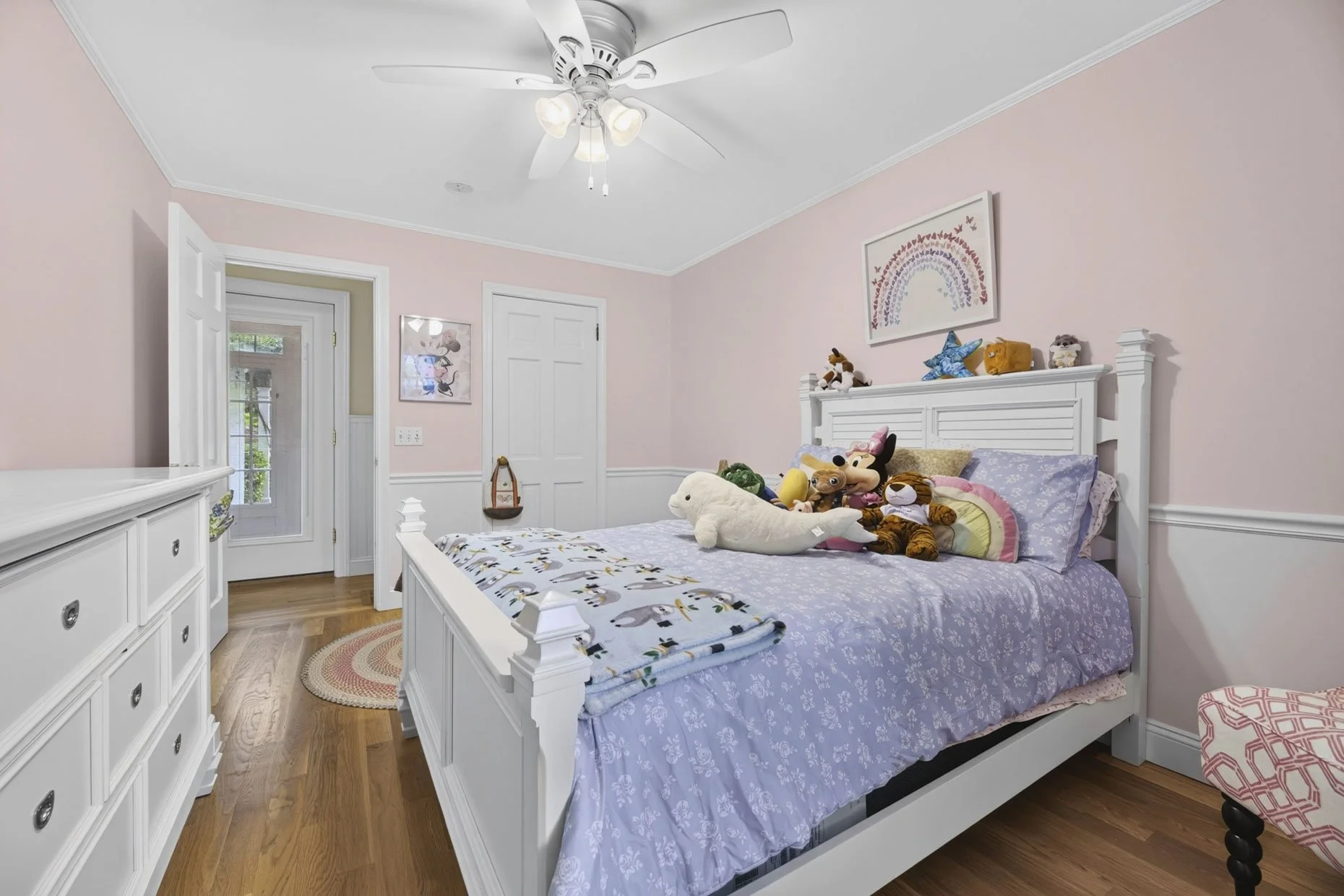 A cozy children's bedroom with pastel pink walls, a white wooden bed, and stuffed animals on the bed including a large white seal, a teddy bear, a lion, and characters resembling Disney's Minnie Mouse. There is a white dresser with six drawers on the