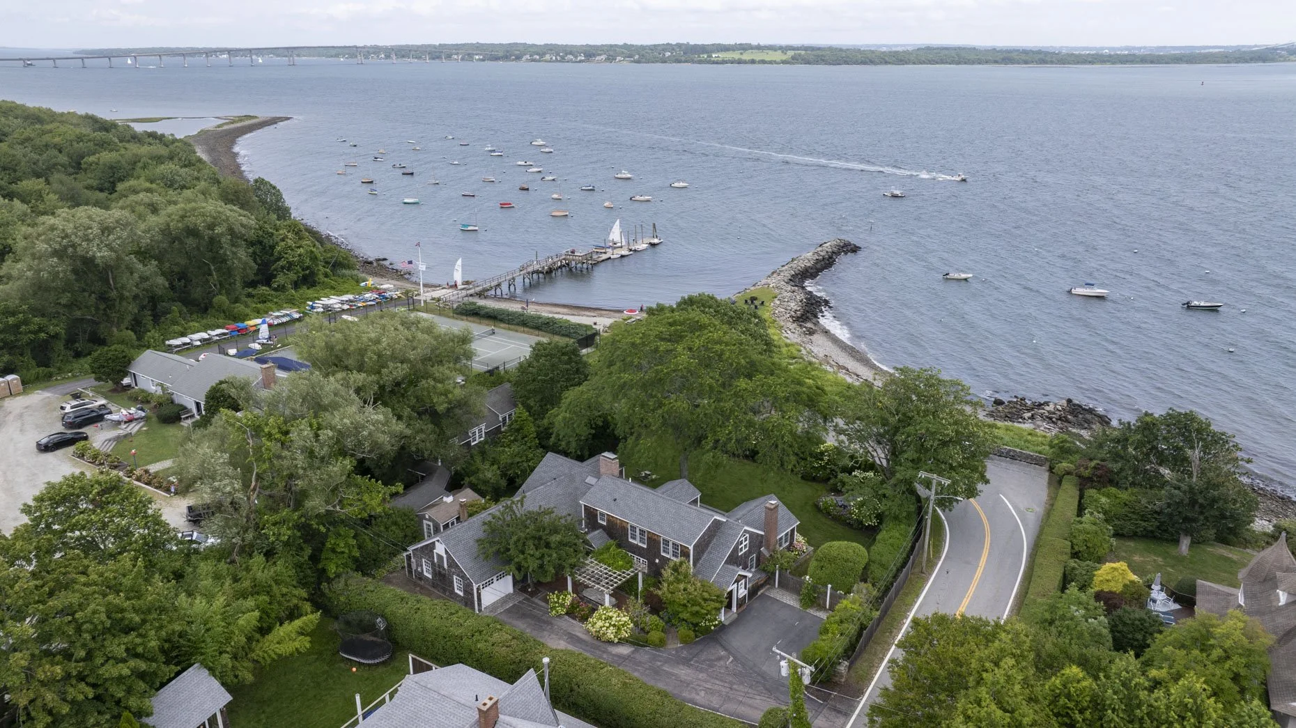 Aerial view of a coastal neighborhood with houses, a tennis court, a marina with boats, and a bridge in the distance over a large body of water.