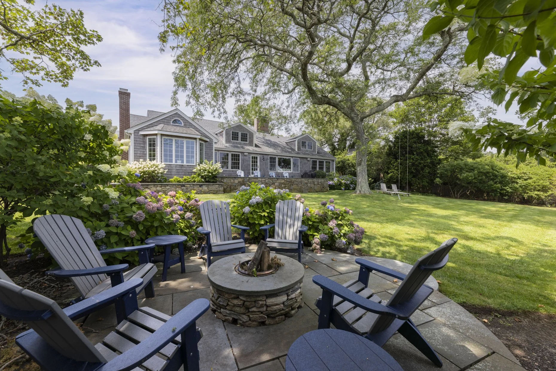 A backyard with a circular patio seating area with Adirondack chairs surrounding a fire pit, lush green grass, flowering bushes, a large tree with branches overhead, and a house with multiple chimneys and large windows in the background