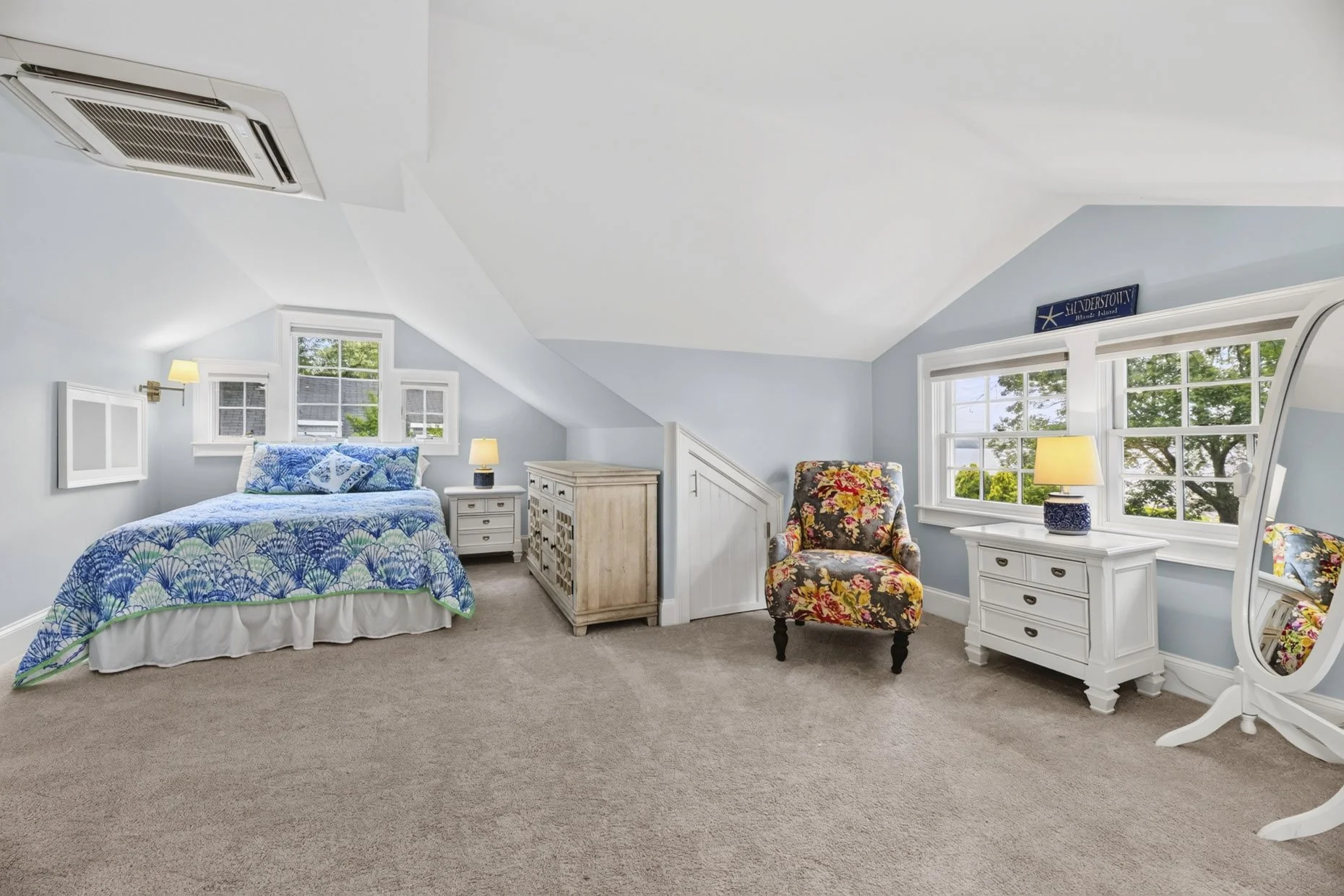 Bright bedroom with a sloped ceiling, featuring a bed with blue and green patterned bedding, white nightstands with lamps, a floral armchair, and windows that show greenery outside.