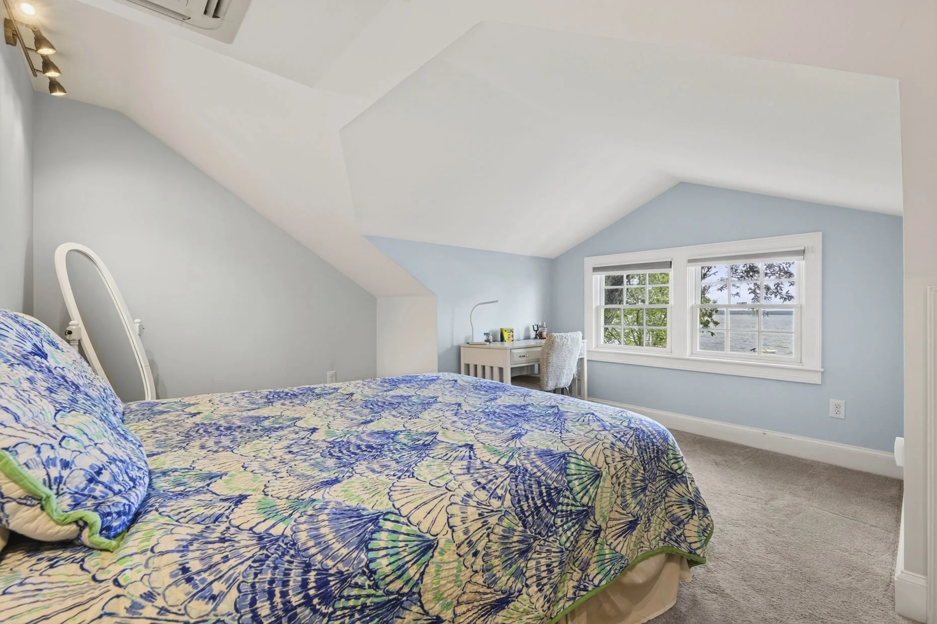 A bedroom with sloped ceiling, light blue walls, and a window showing trees outside. There is a bed with a blue and green butterfly patterned comforter and pillows. A small white desk with a chair is positioned beneath the window, with some items on 