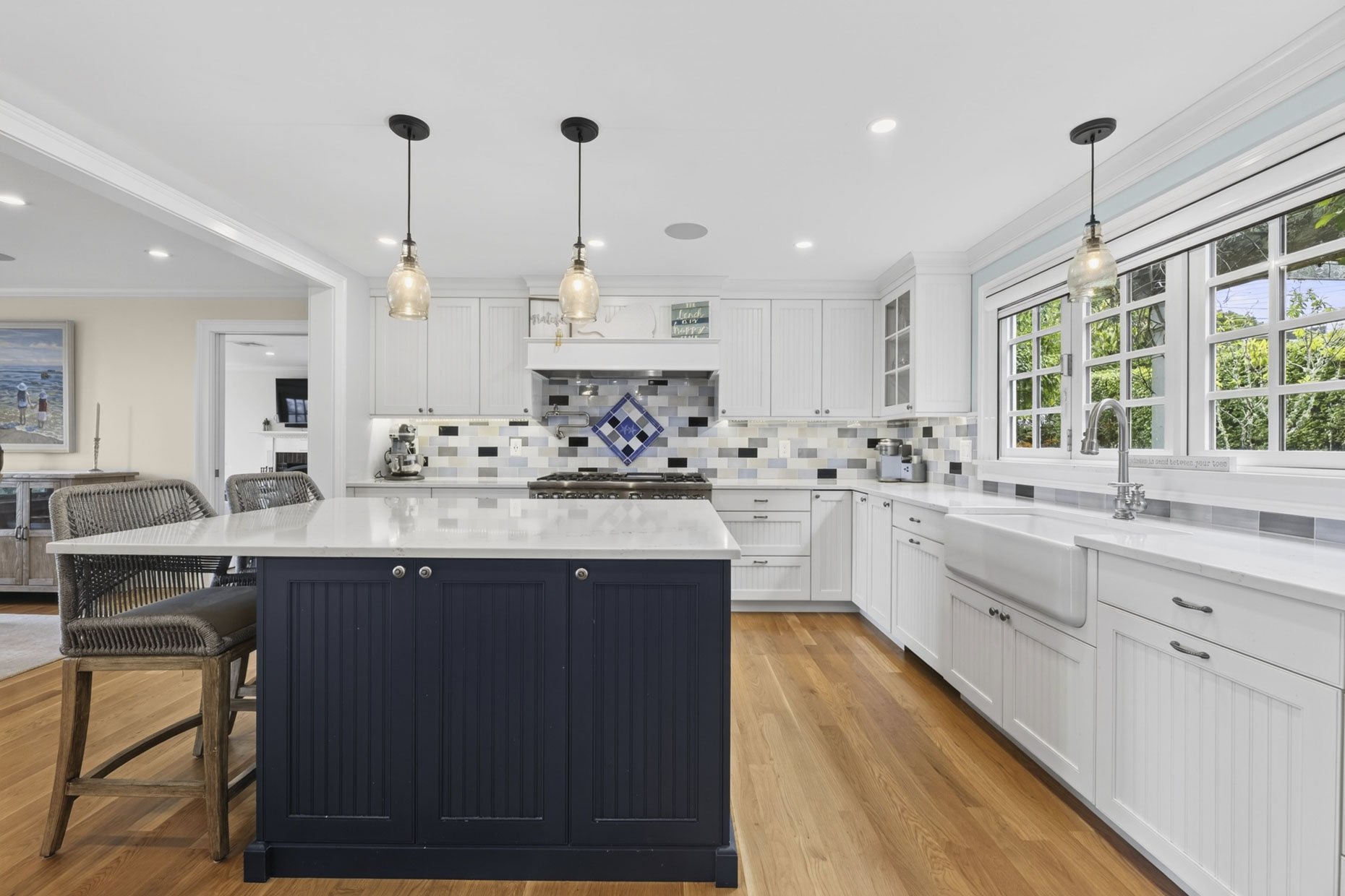 Kitchen with white cabinets, a navy blue island, wooden floors, pendant lights, and large windows overlooking greenery.