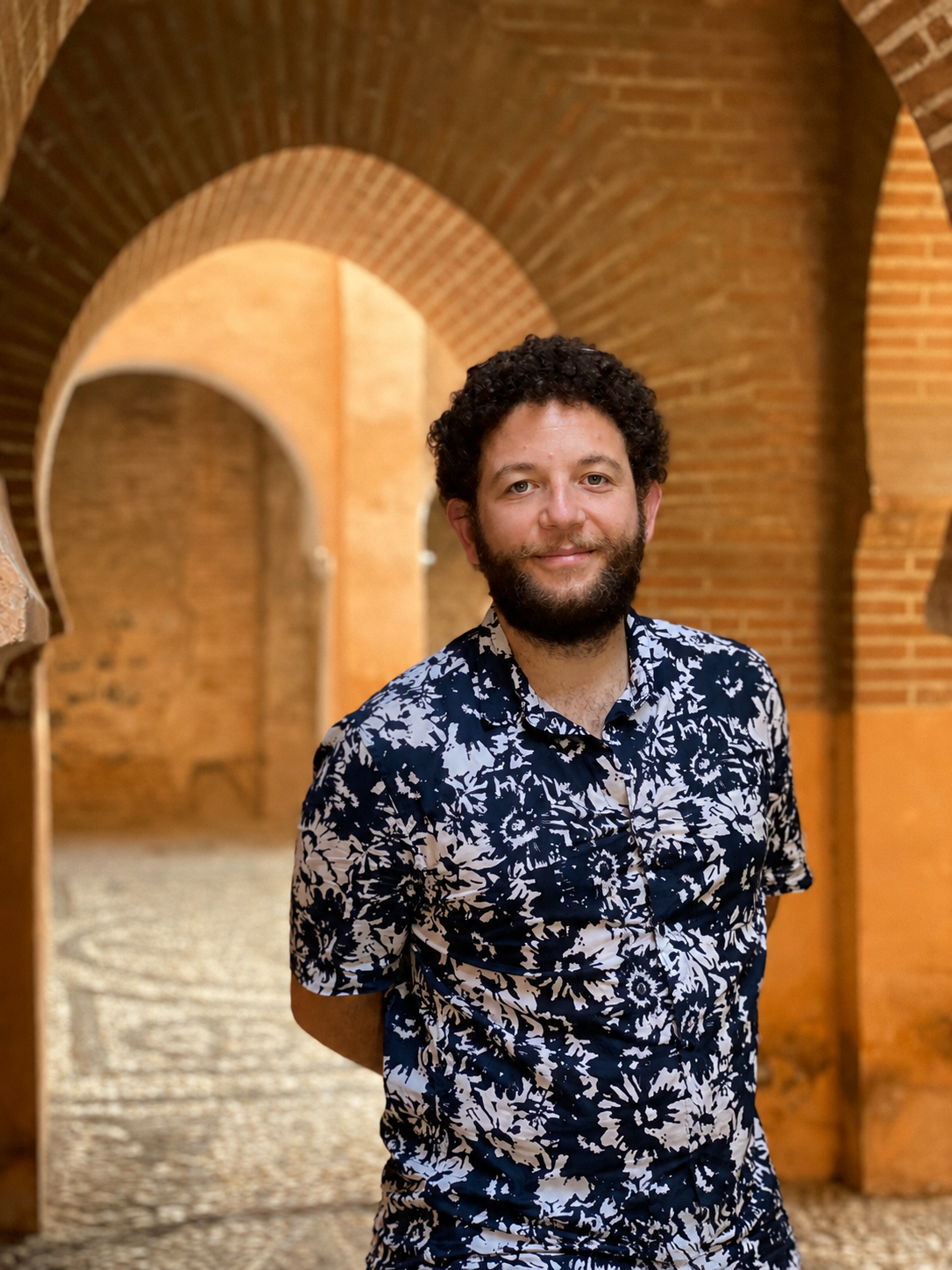 A man with curly hair, beard, and wearing a black and white patterned shirt standing under brick arches.