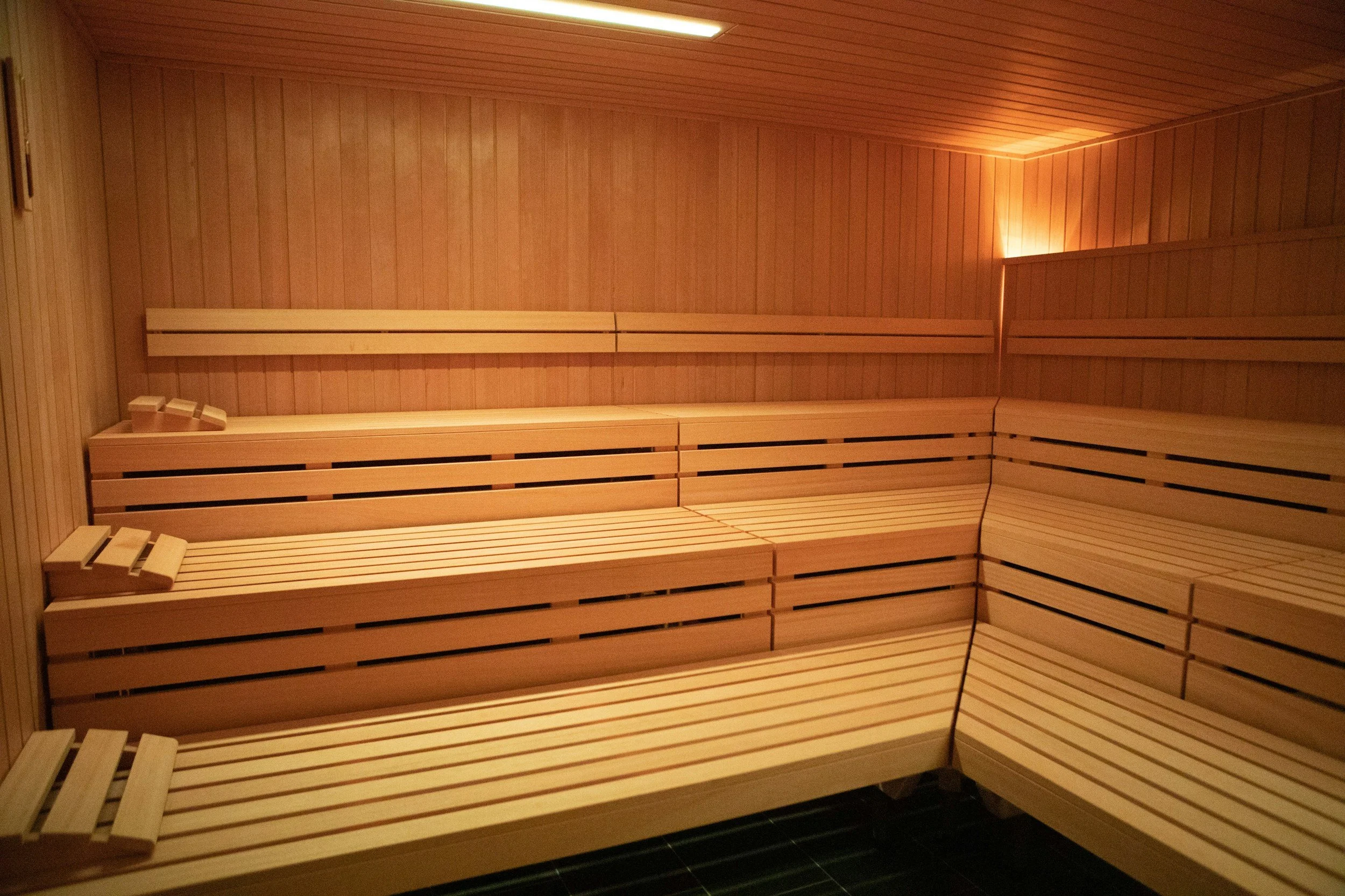 Ein Holzsauna-Innenraum mit mehreren Sitzbänken aus hellem Holz und einer abgehängten Decke mit indirekter Beleuchtung.