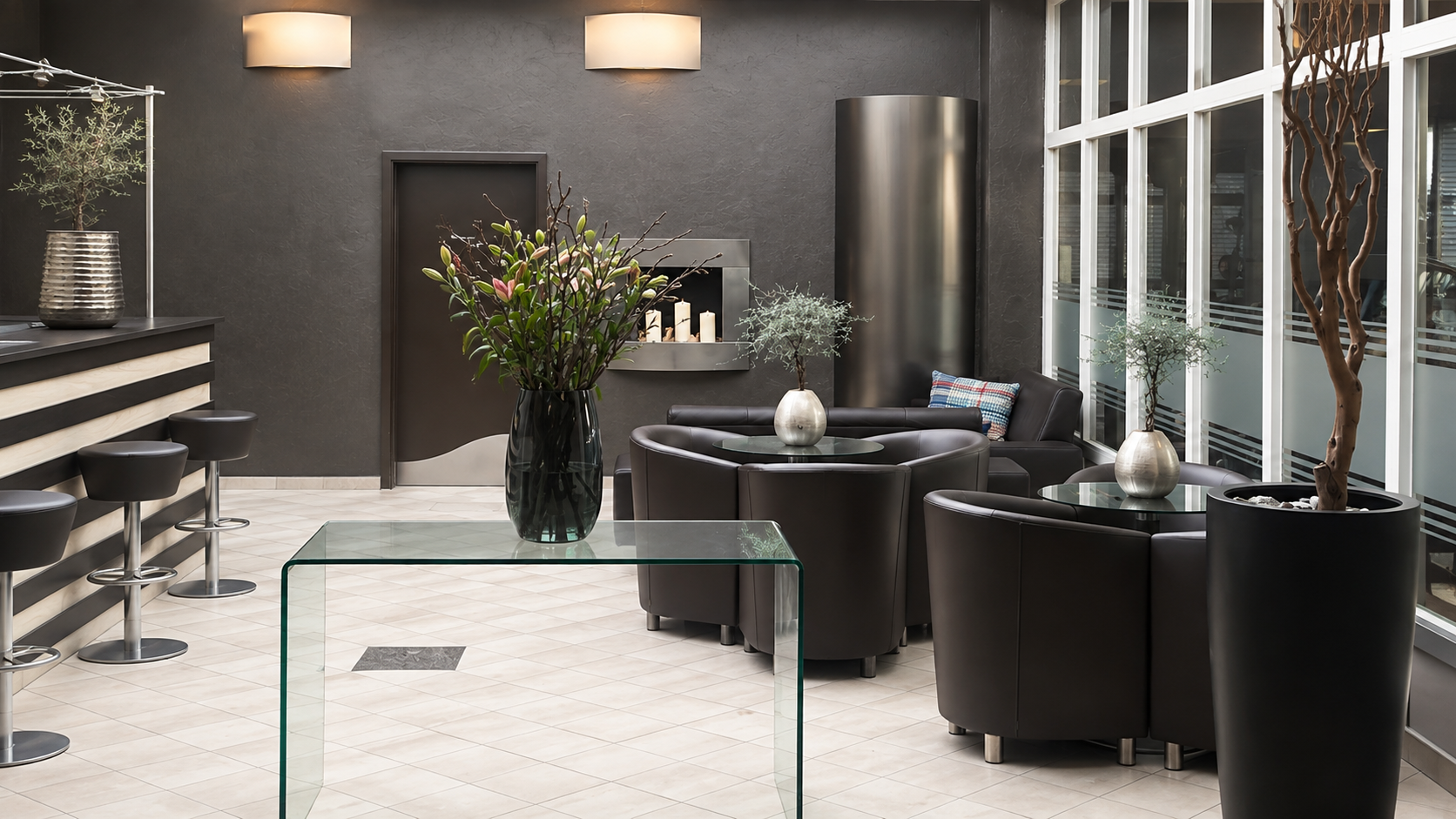 Moderne Hotel-Lounge mit schwarzen Sesseln, großen Vasen mit Bäumen und Blick auf die große Fensterfront.